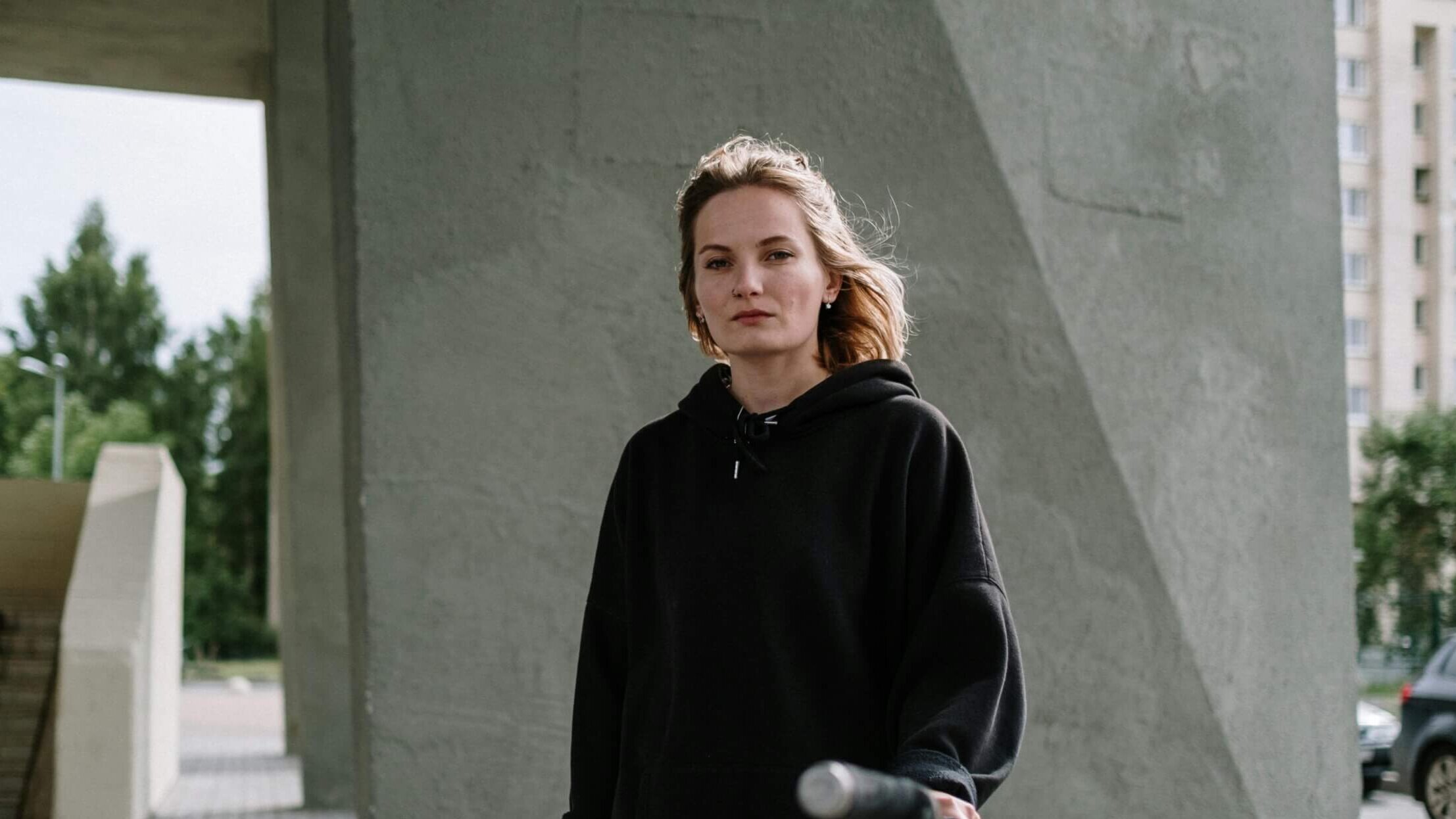 Young woman in a black hoodie standing outdoors holding a scooter handlebar near a concrete structure