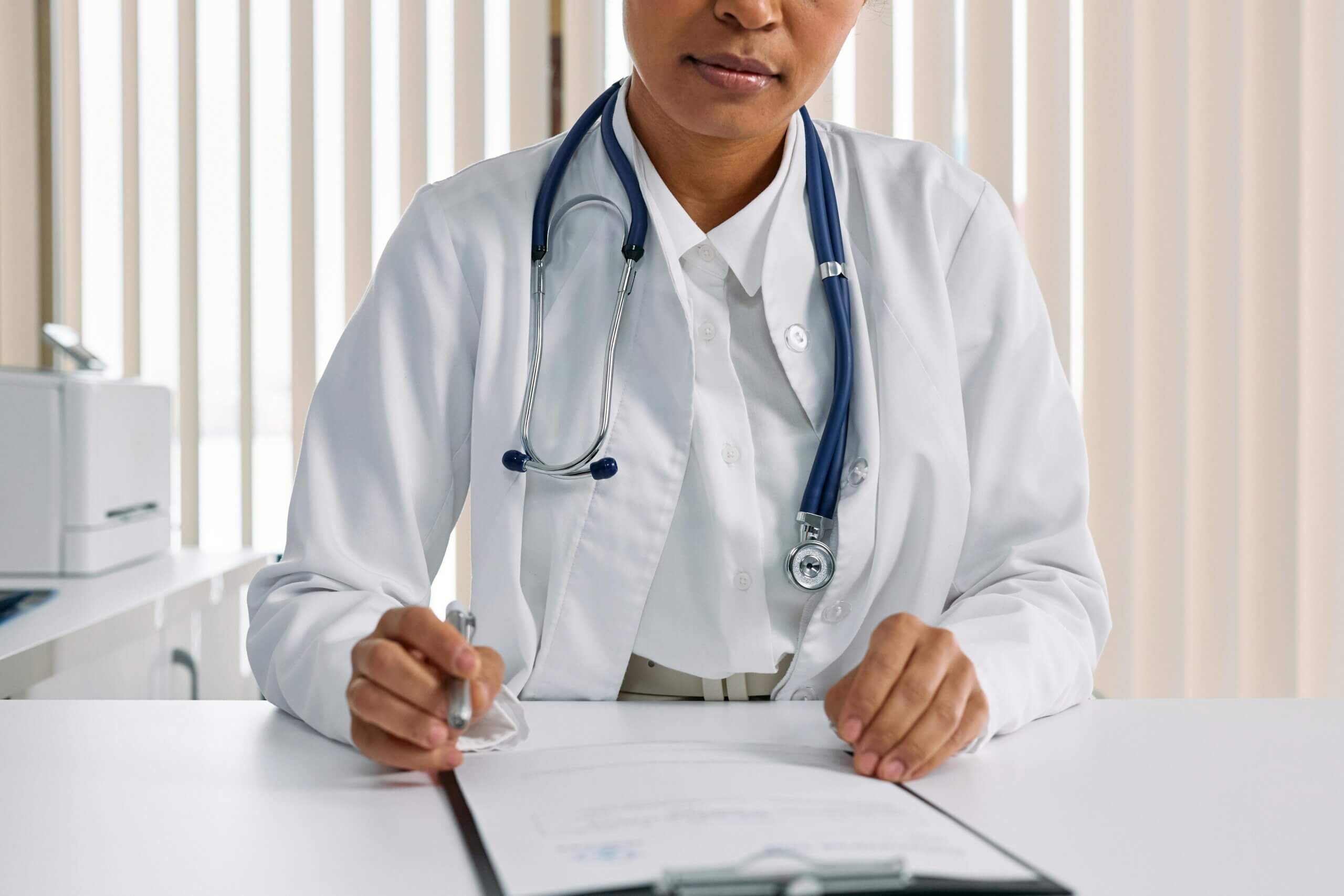 Doctor in white coat with stethoscope writing on a clipboard at a desk