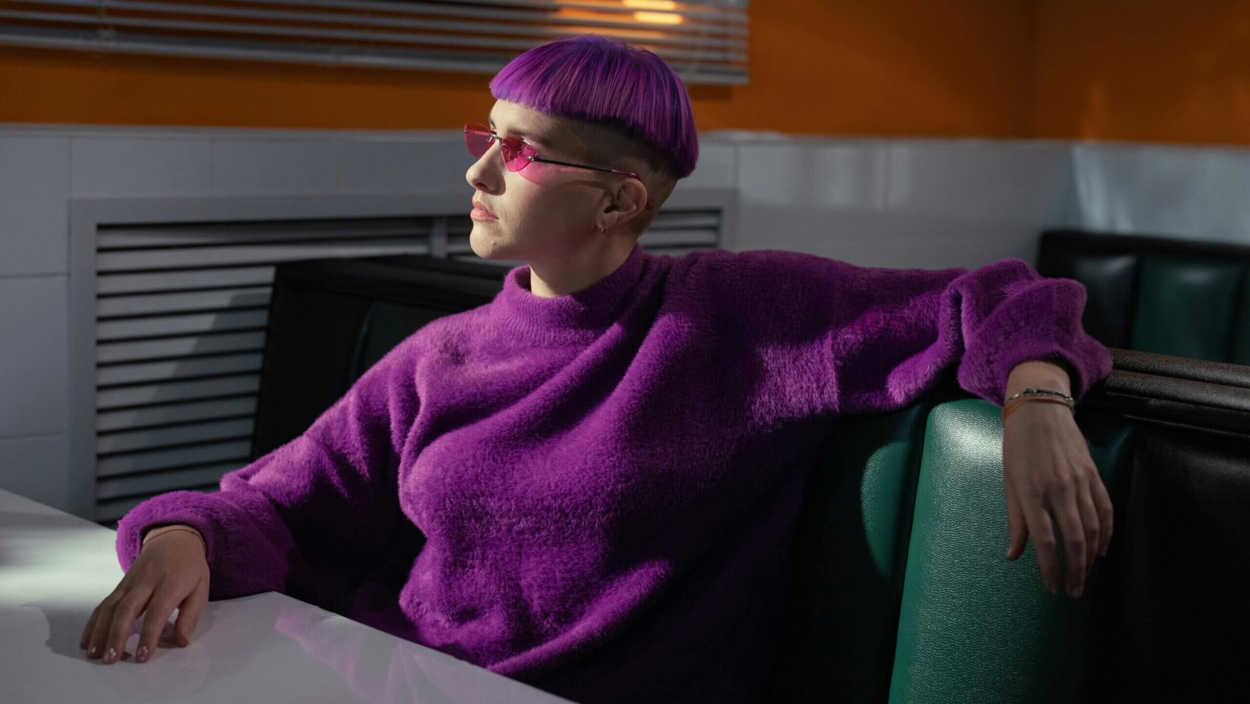Person with purple bowl-cut hair and pink sunglasses wearing a purple sweater sitting in a diner booth