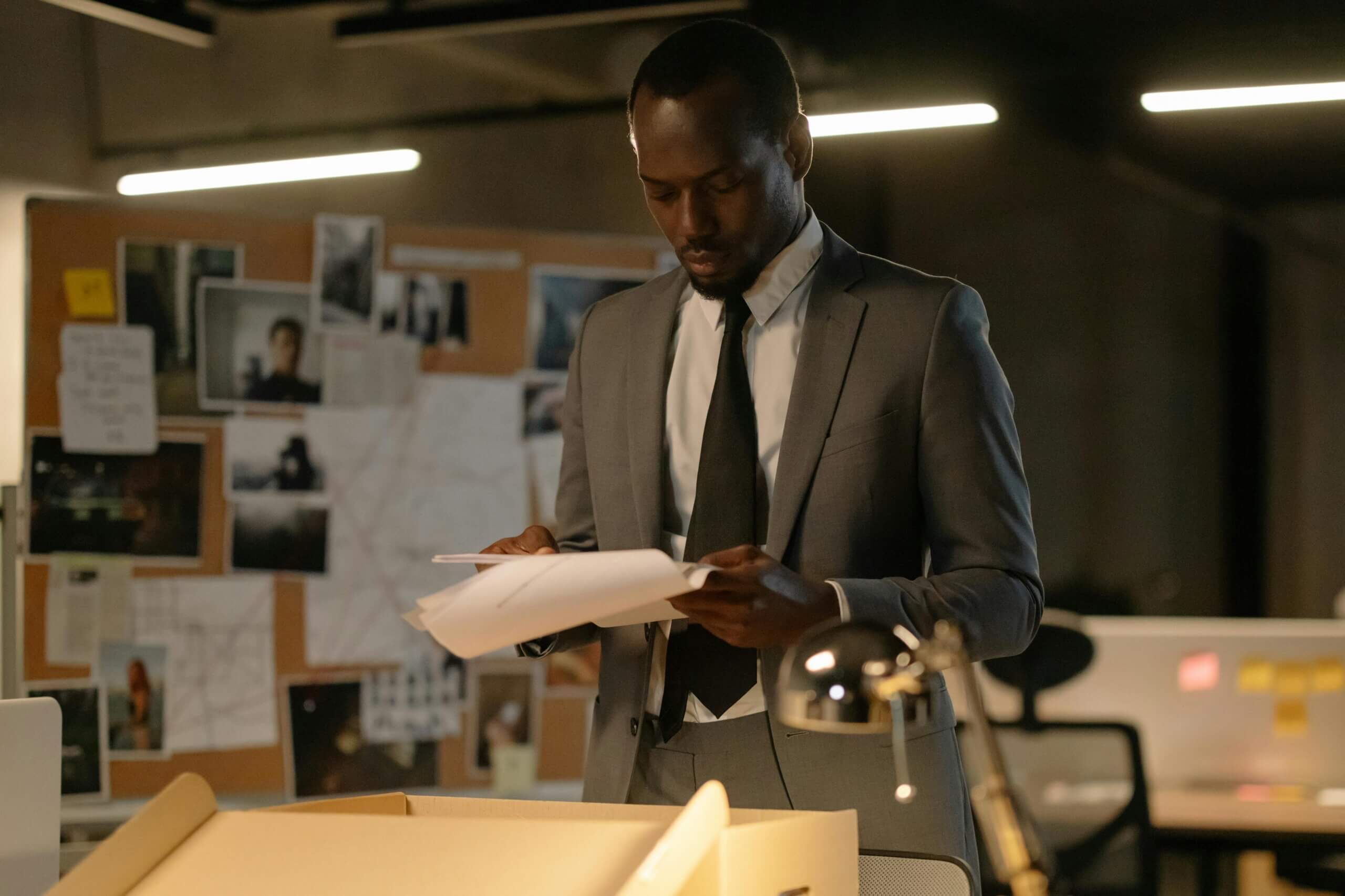 Man in gray suit and black tie reviewing documents in an office with a bulletin board covered in photos and notes.