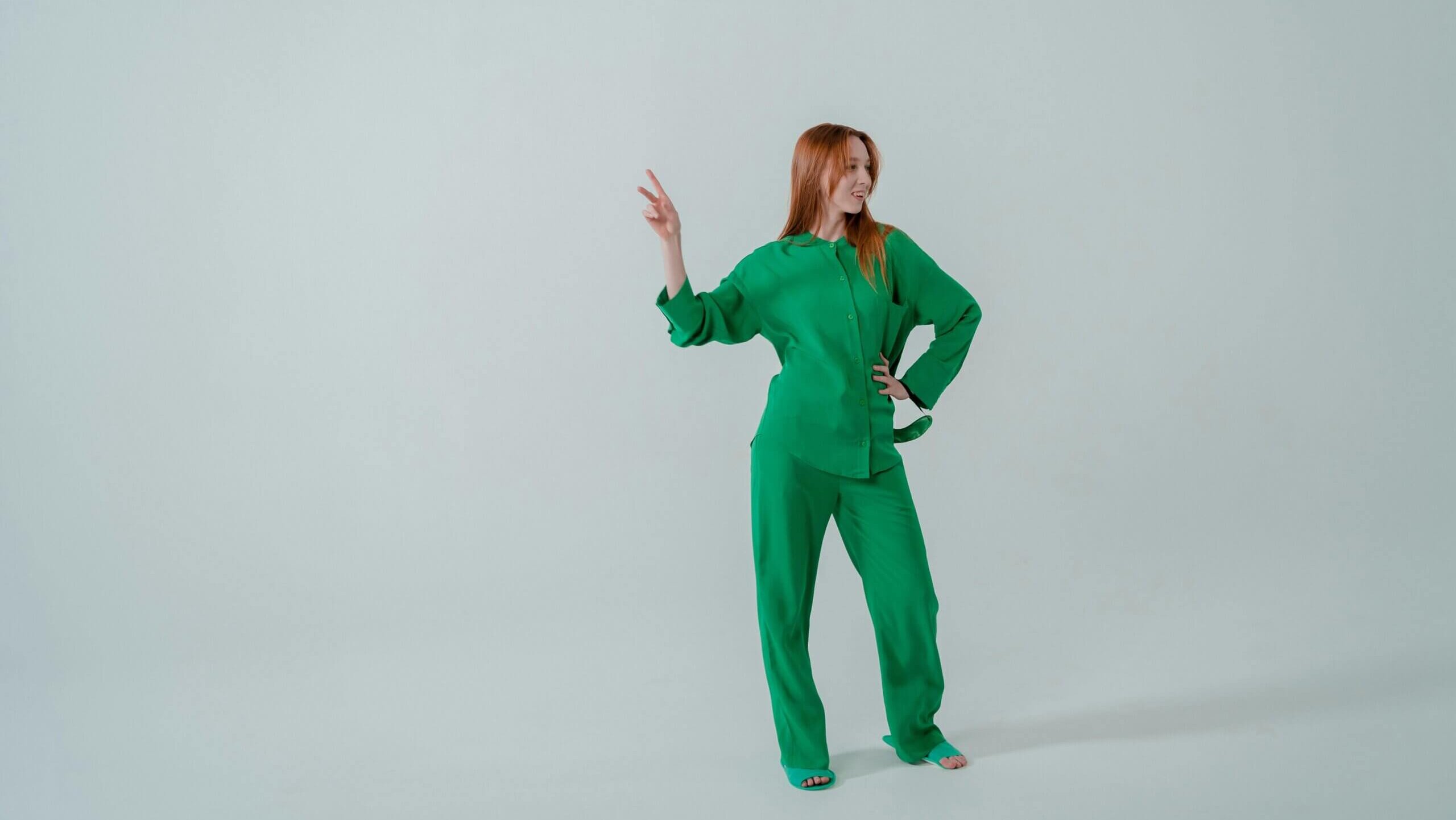 Woman in green outfit making a peace sign with her right hand against a plain light background