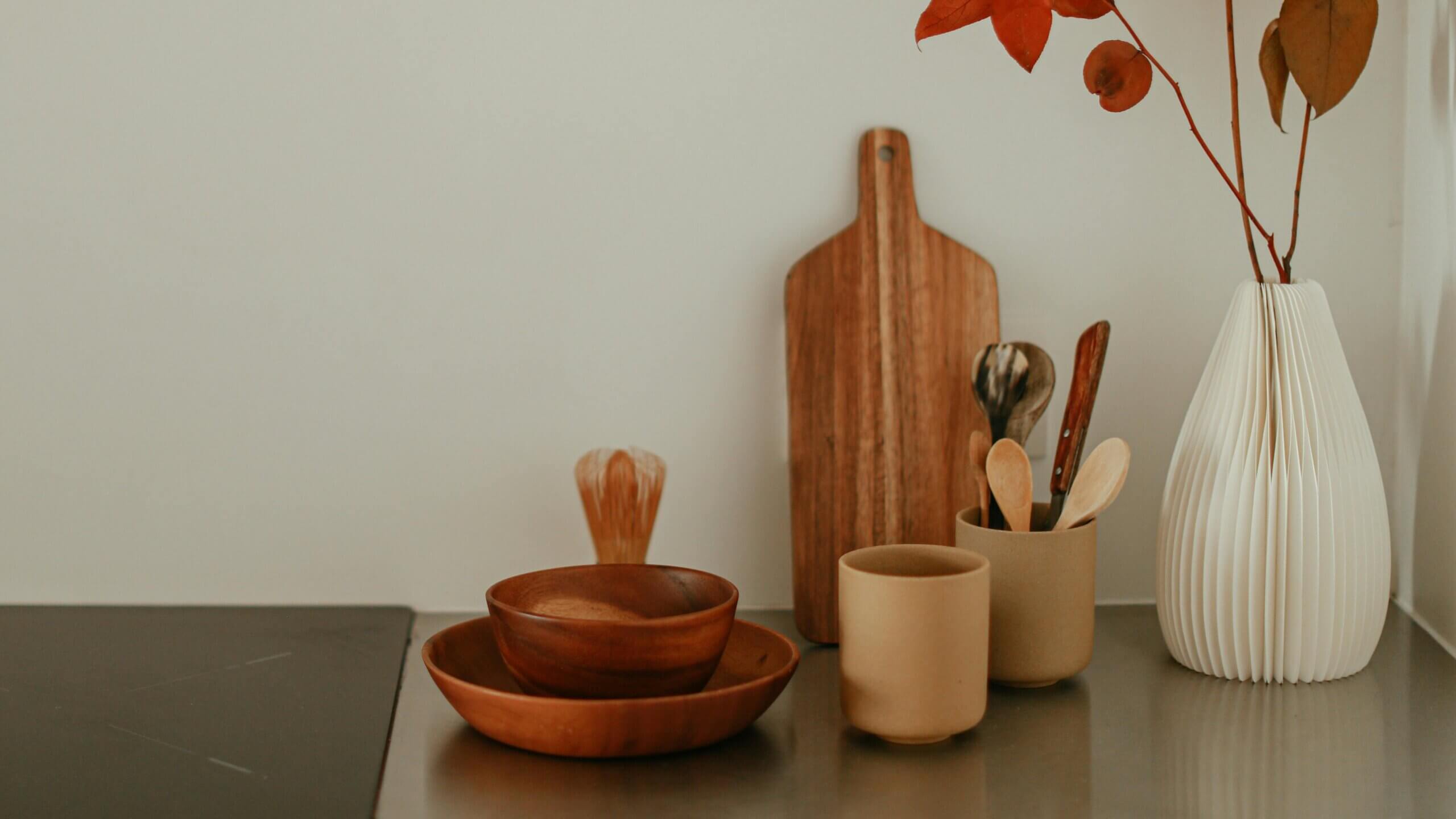 Wooden bowls, ceramic cups, utensils, cutting board, and white vase with autumn leaves on kitchen counter