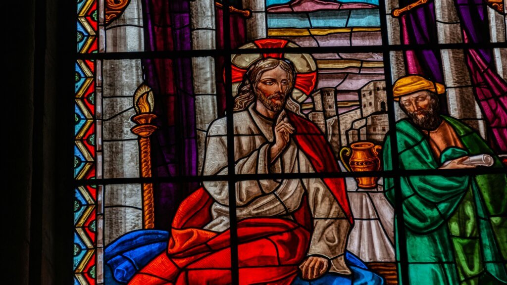 Stained glass window depicting Jesus with a halo and red robe, seated beside a man in green holding a scroll.