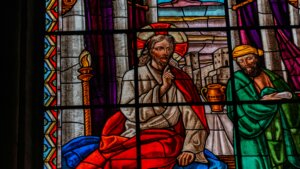 Stained glass window depicting Jesus with a halo and red robe, seated beside a man in green holding a scroll.