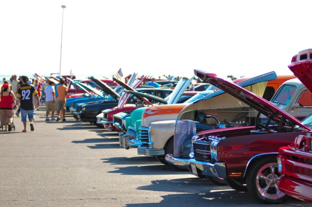 Row of classic cars with open hoods at an outdoor car show with people walking nearby