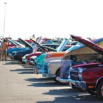 Row of classic cars with open hoods at an outdoor car show with people walking nearby