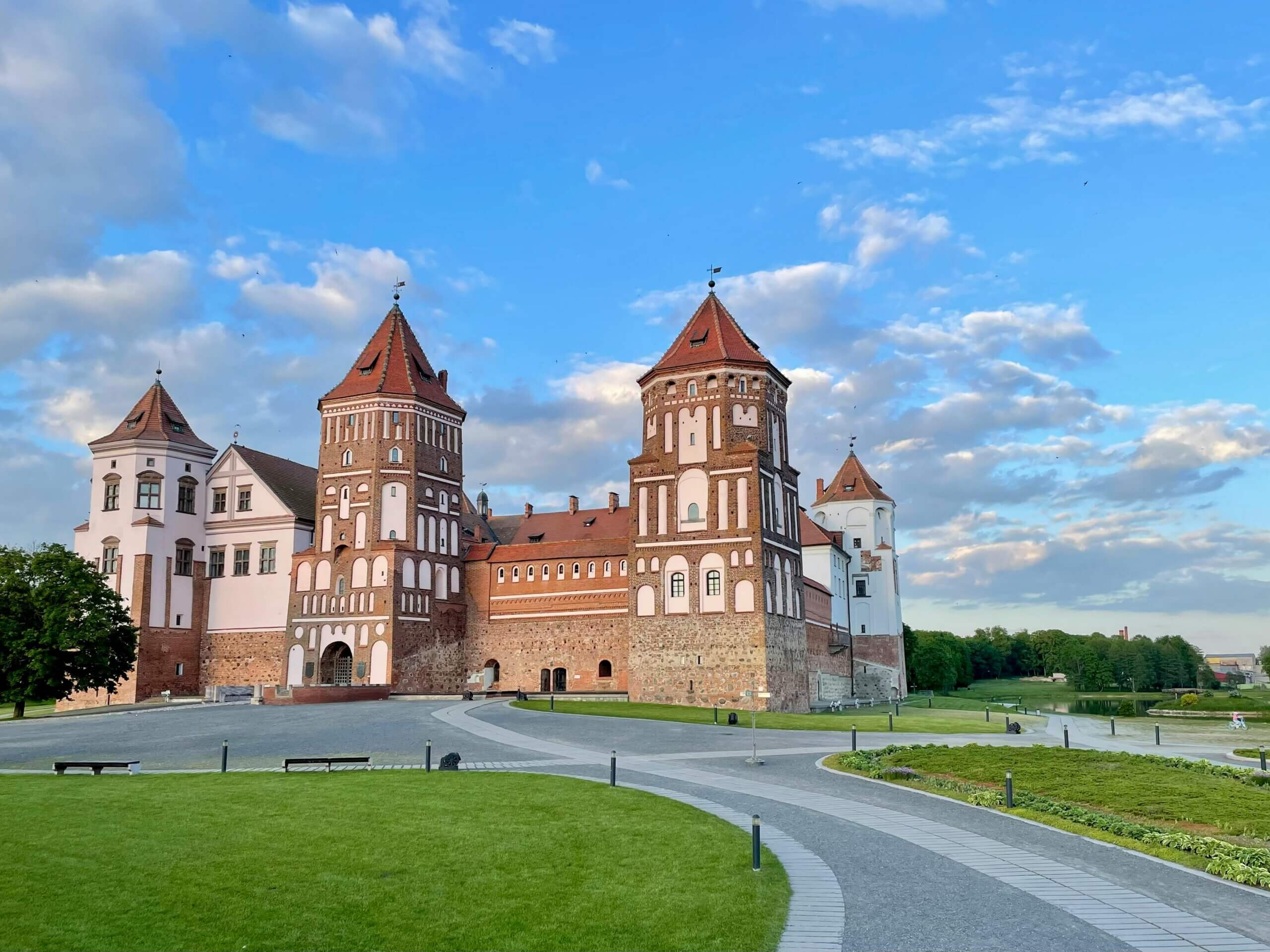 Mir Castle, a historic brick and white stone fortress with red roofs, under a blue sky with scattered clouds.