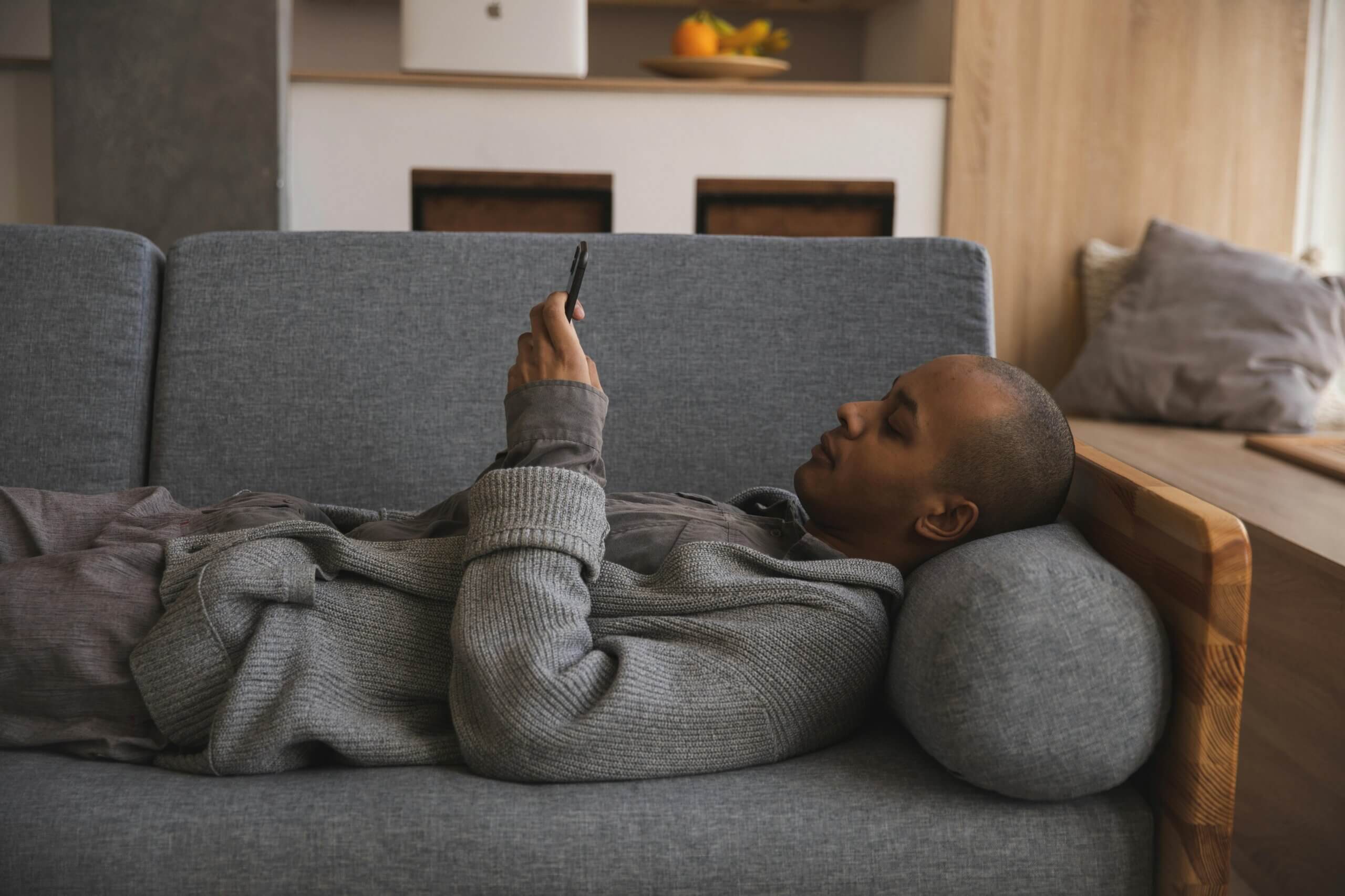 Man lying on gray couch using smartphone, wearing gray sweater and brown shirt.