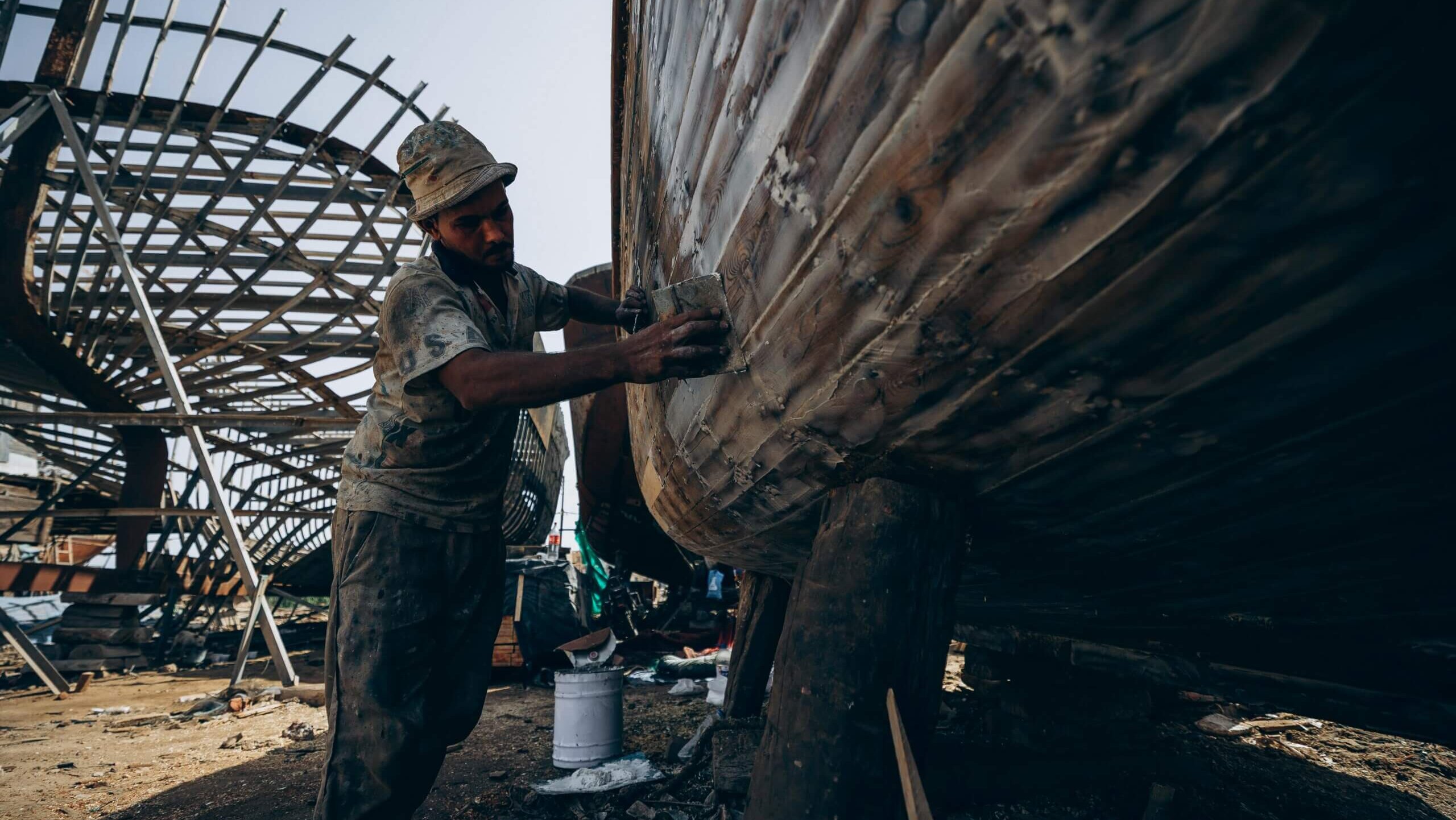 Man sanding the wooden hull of a boat in a shipyard with boat frames in the background