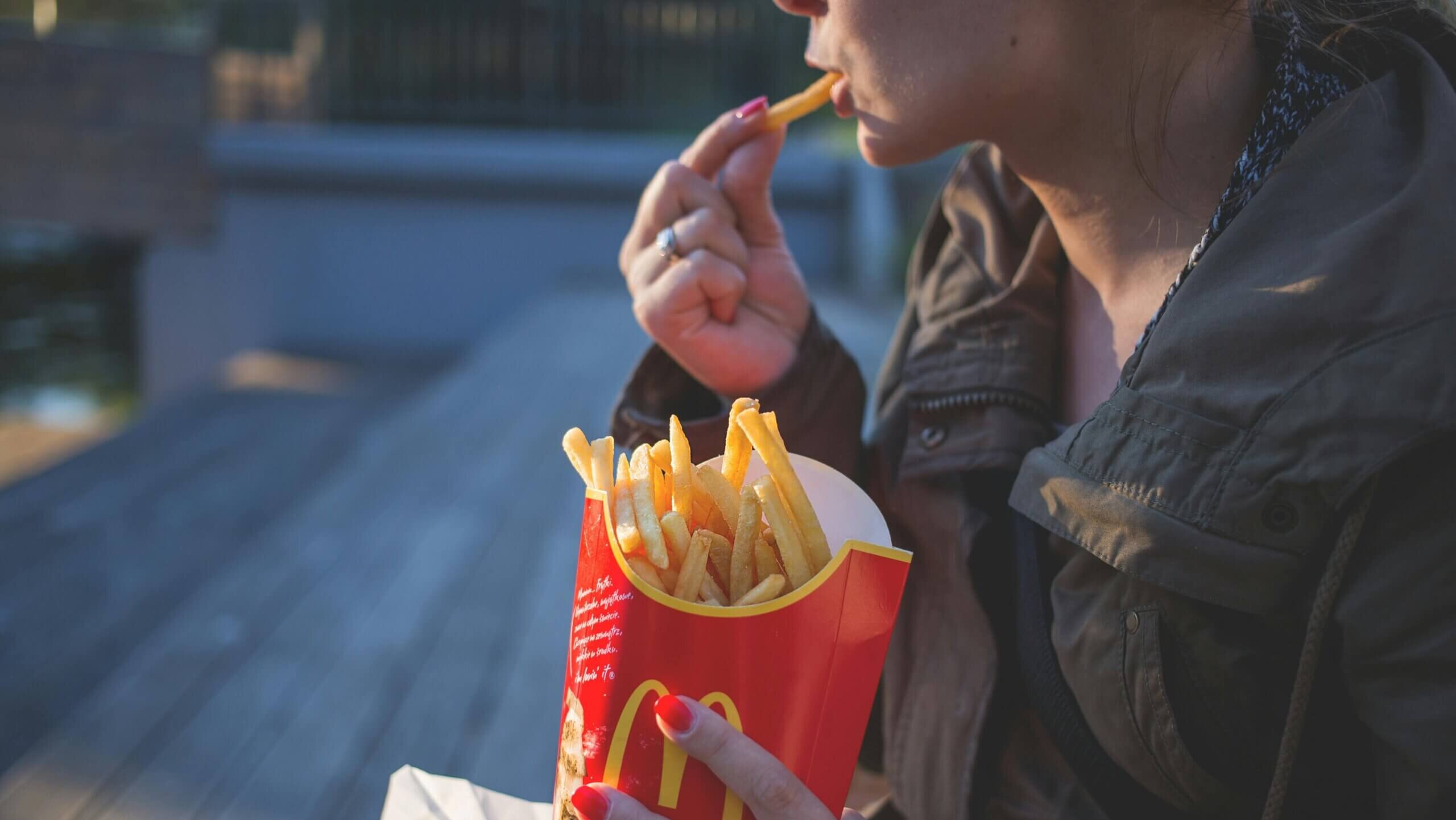 Person eating McDonald's French fries outdoors, wearing a dark jacket and red nail polish.