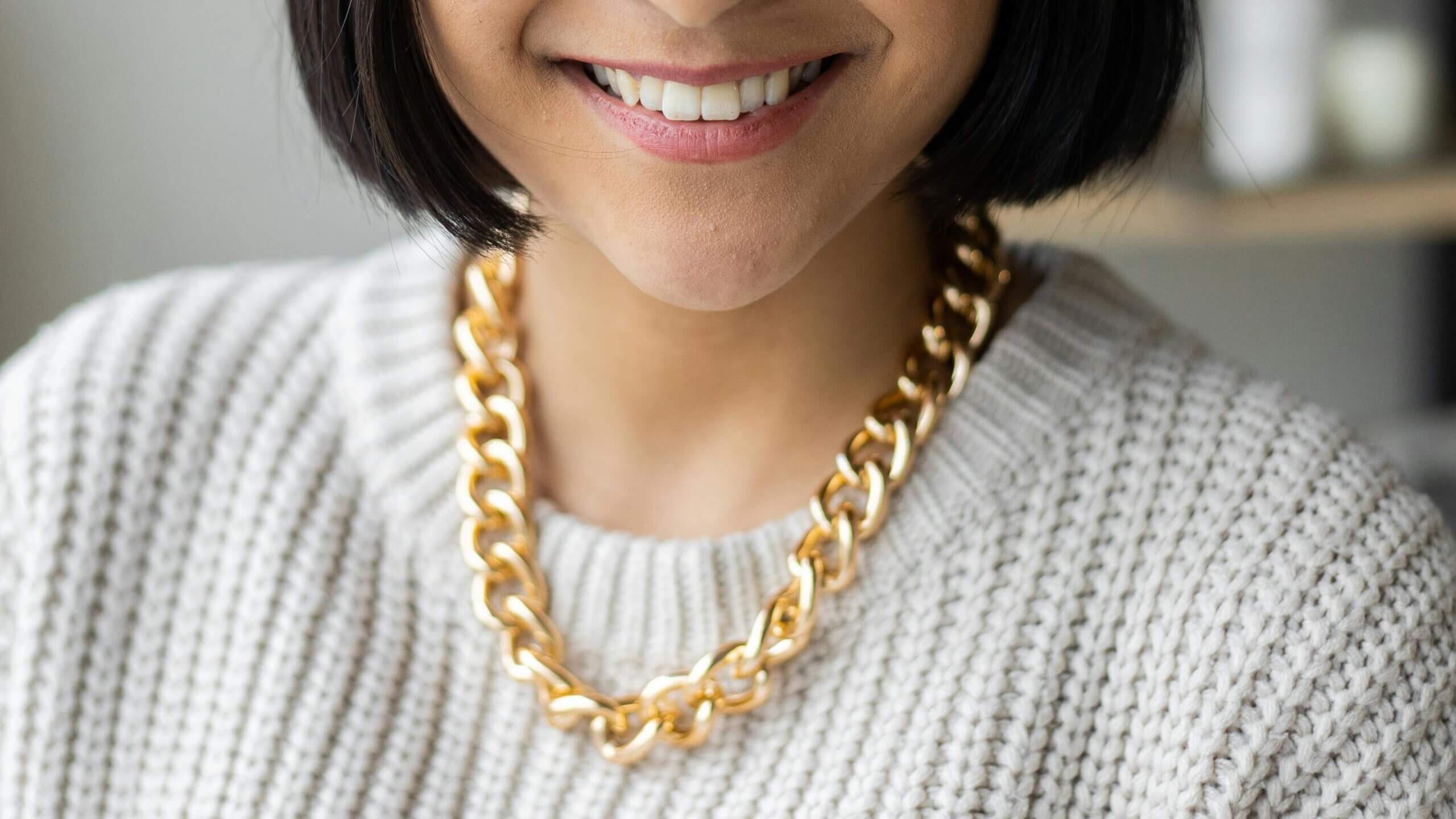 Close-up of a smiling woman wearing a chunky gold chain necklace and a white knitted sweater.