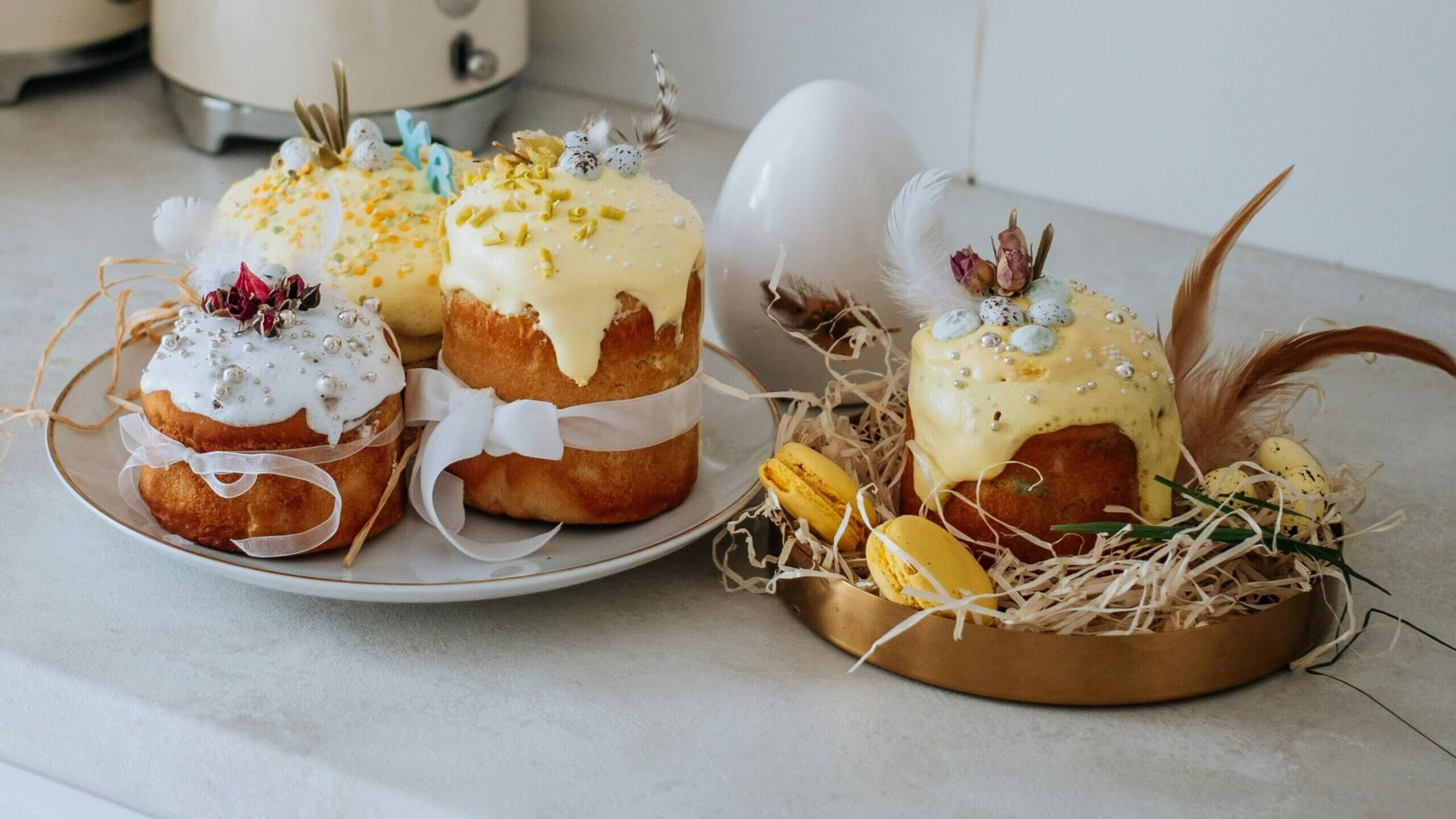 Easter cakes with icing, decorated with feathers, small eggs, and macarons on a kitchen counter.