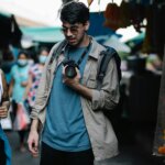 Two young adults wearing sunglasses walk through a market, one holding a camera and the other with a blue jacket tied around the waist.