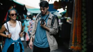 Two young adults wearing sunglasses walk through a market, one holding a camera and the other with a blue jacket tied around the waist.