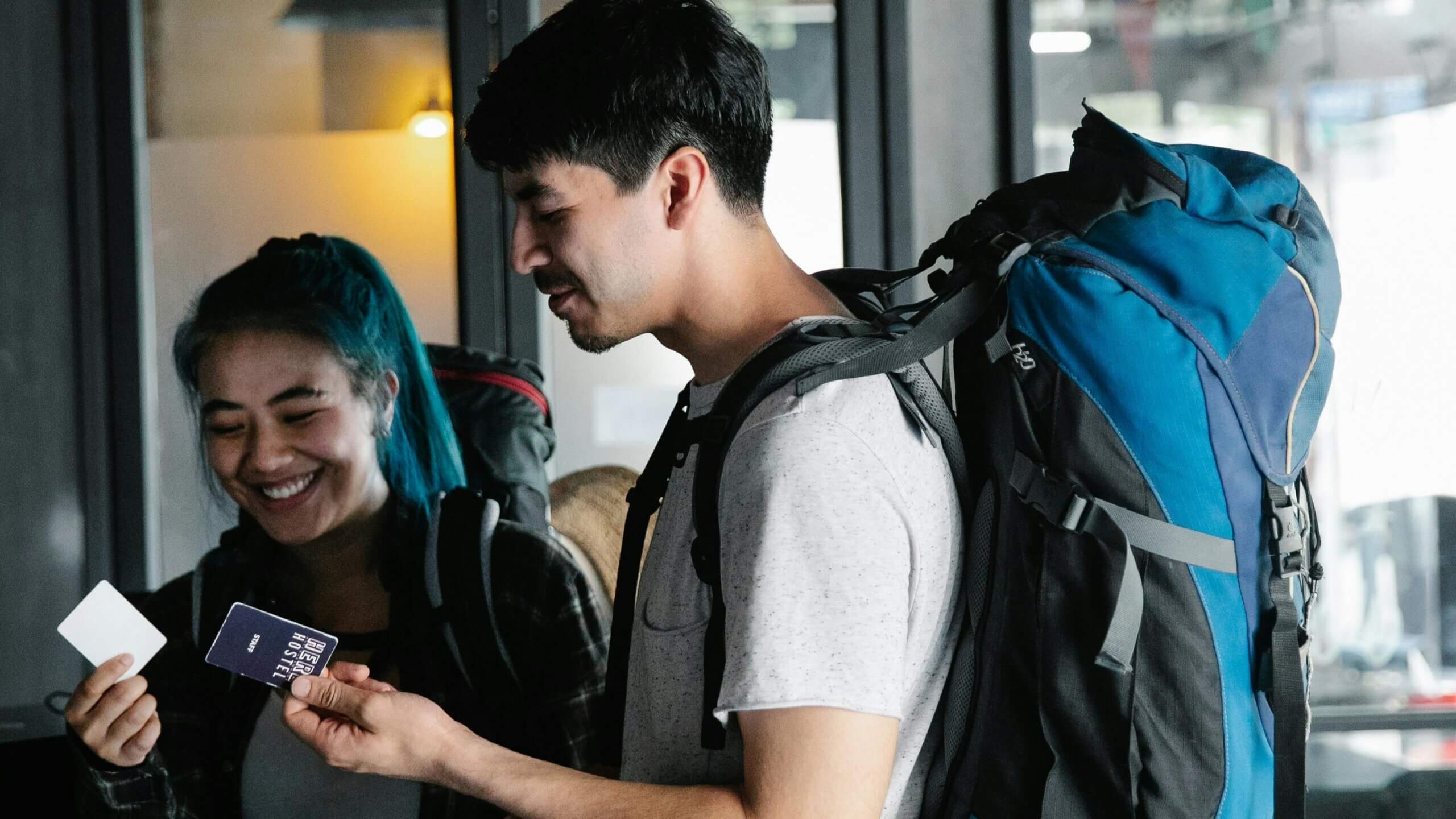Two backpackers smiling and holding hostel key cards inside a building.