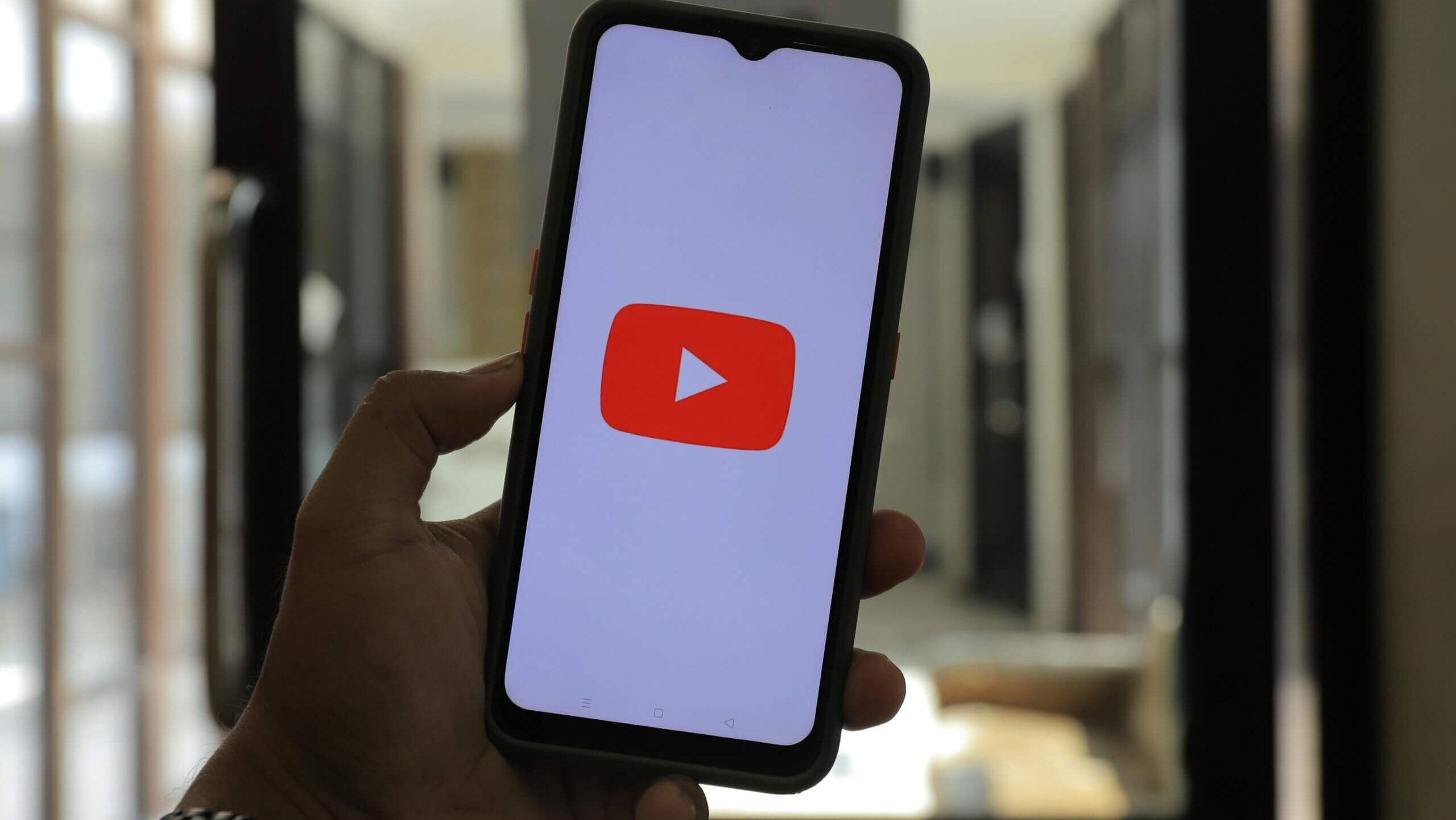 Hand holding smartphone displaying the YouTube logo on screen in a blurred indoor setting