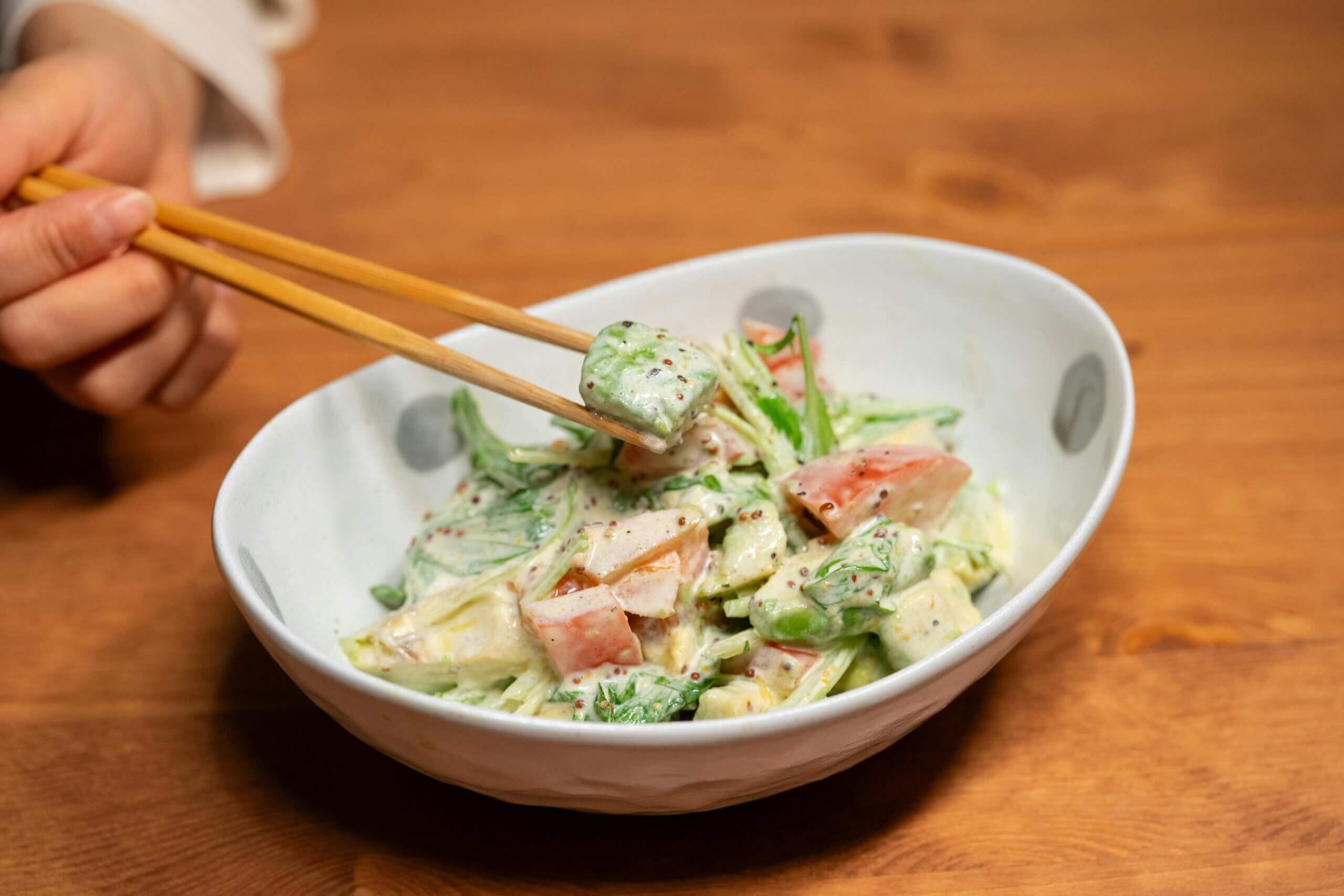 Hand holding chopsticks picking up a piece of creamy salad with vegetables in a white bowl on a wooden table