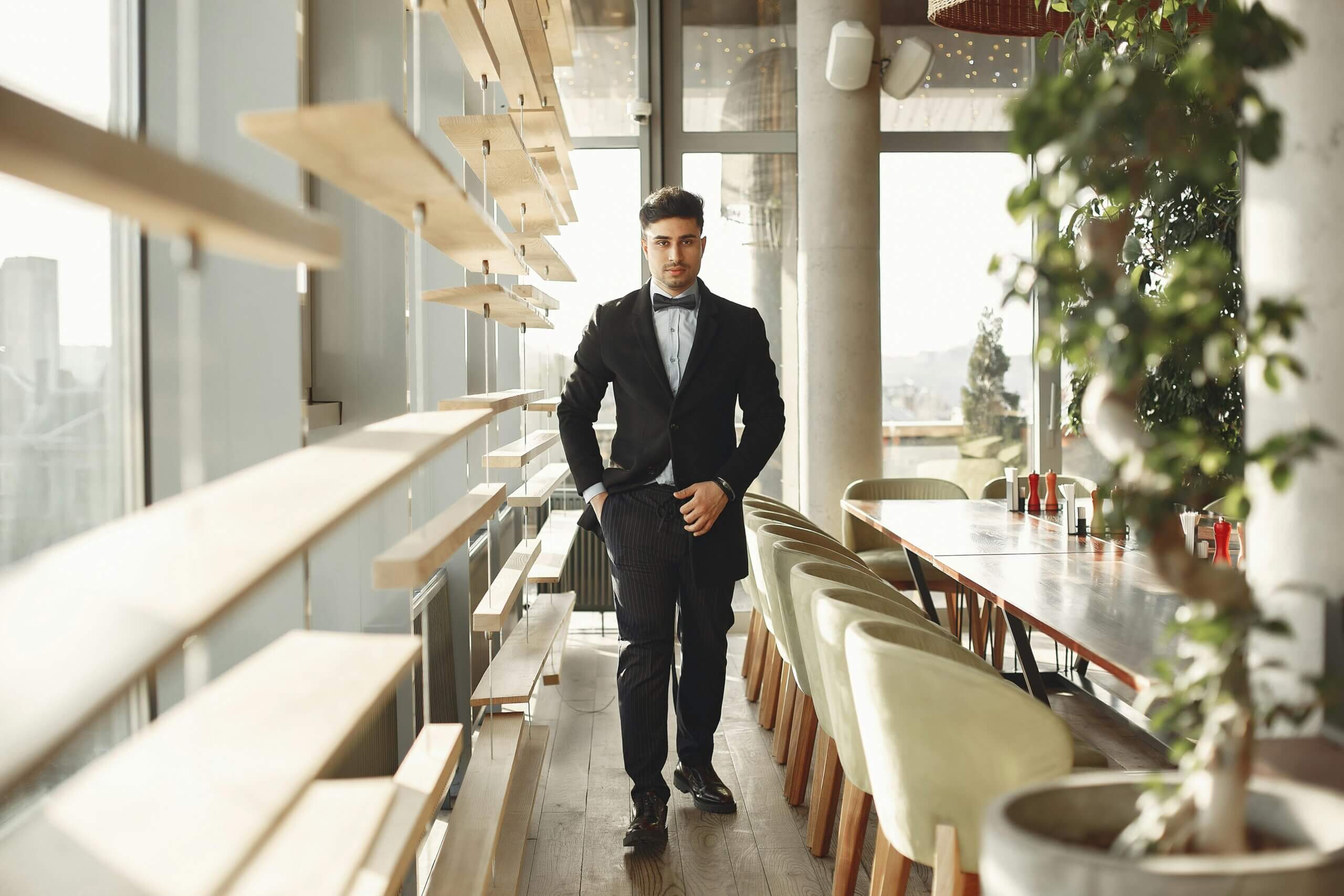 Man in black tuxedo walking in a modern restaurant with wooden shelves and green chairs by a long table