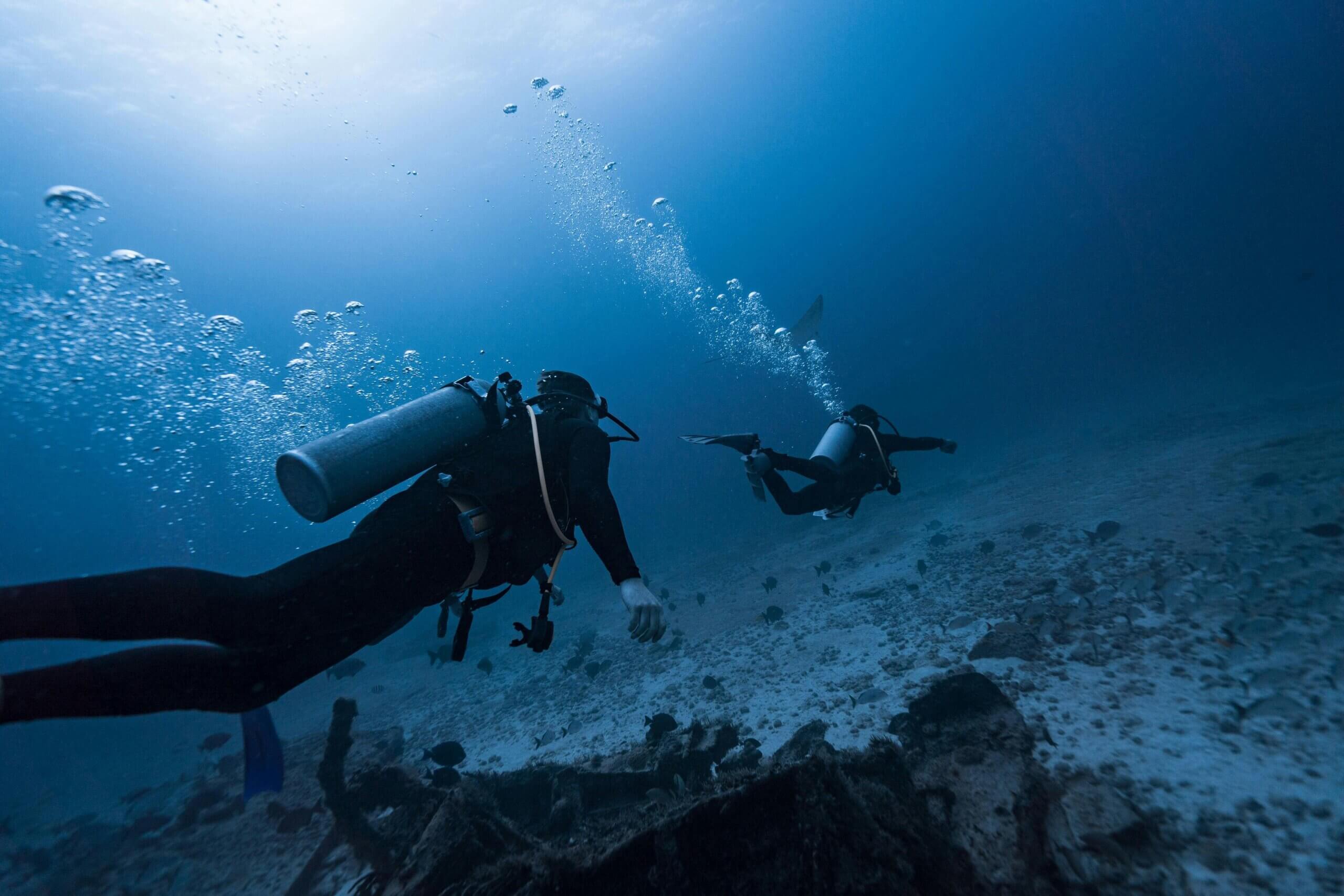Two scuba divers exploring an underwater wreck with fish swimming around in deep blue water.