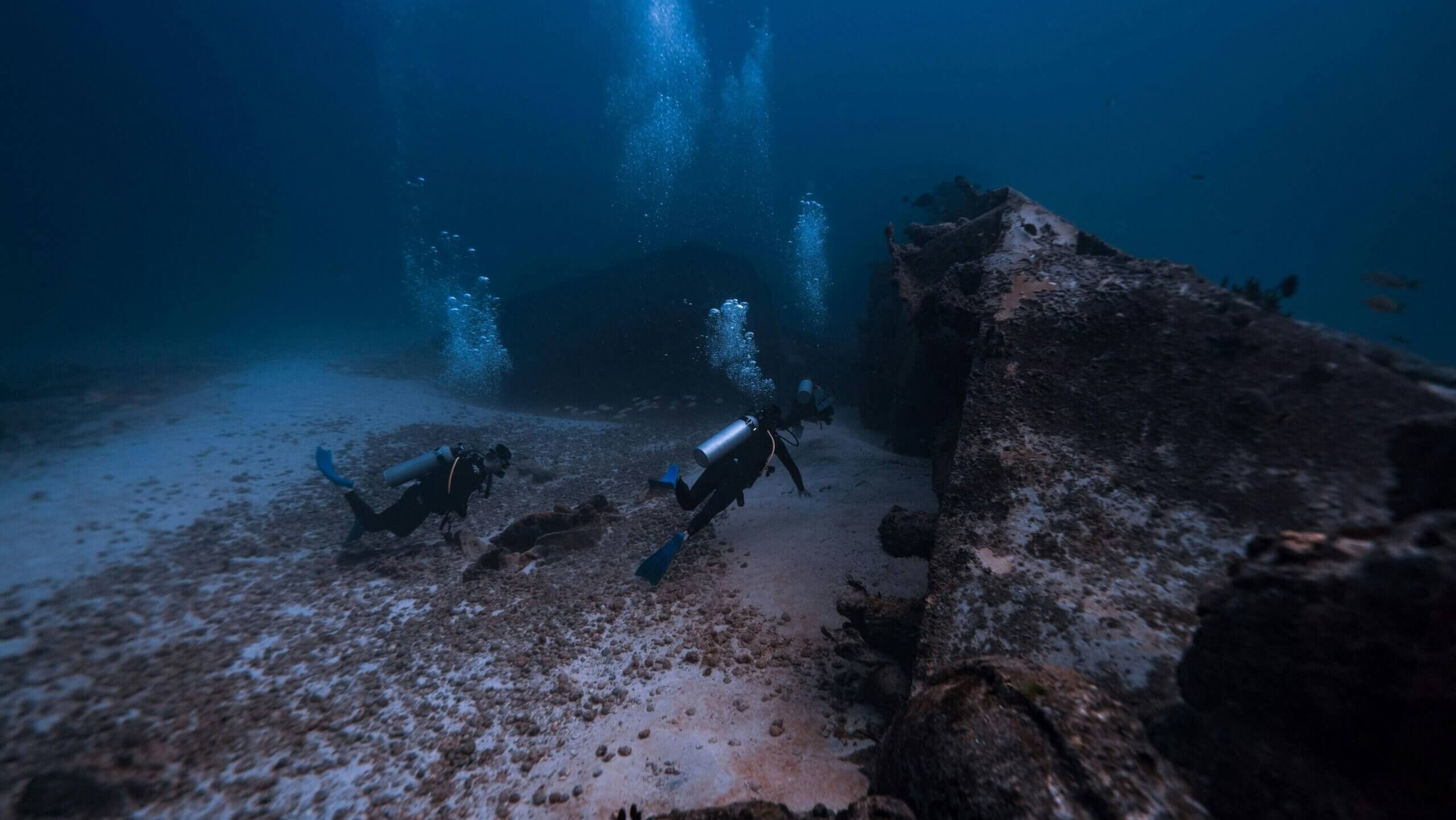 Two scuba divers exploring a deep underwater shipwreck surrounded by marine life and bubbles.
