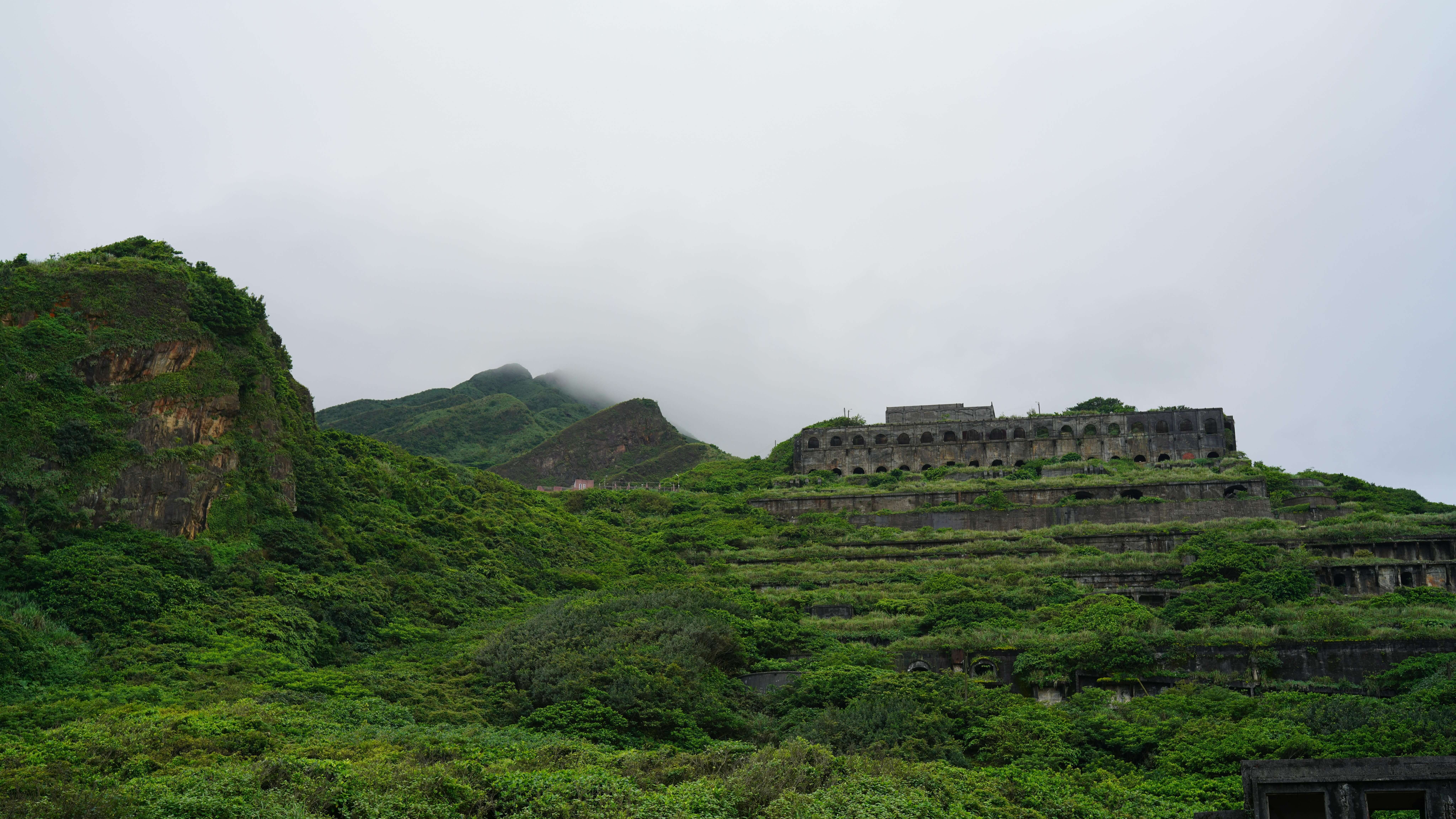 Overgrown terraced hillside with a large abandoned concrete structure under a cloudy sky.