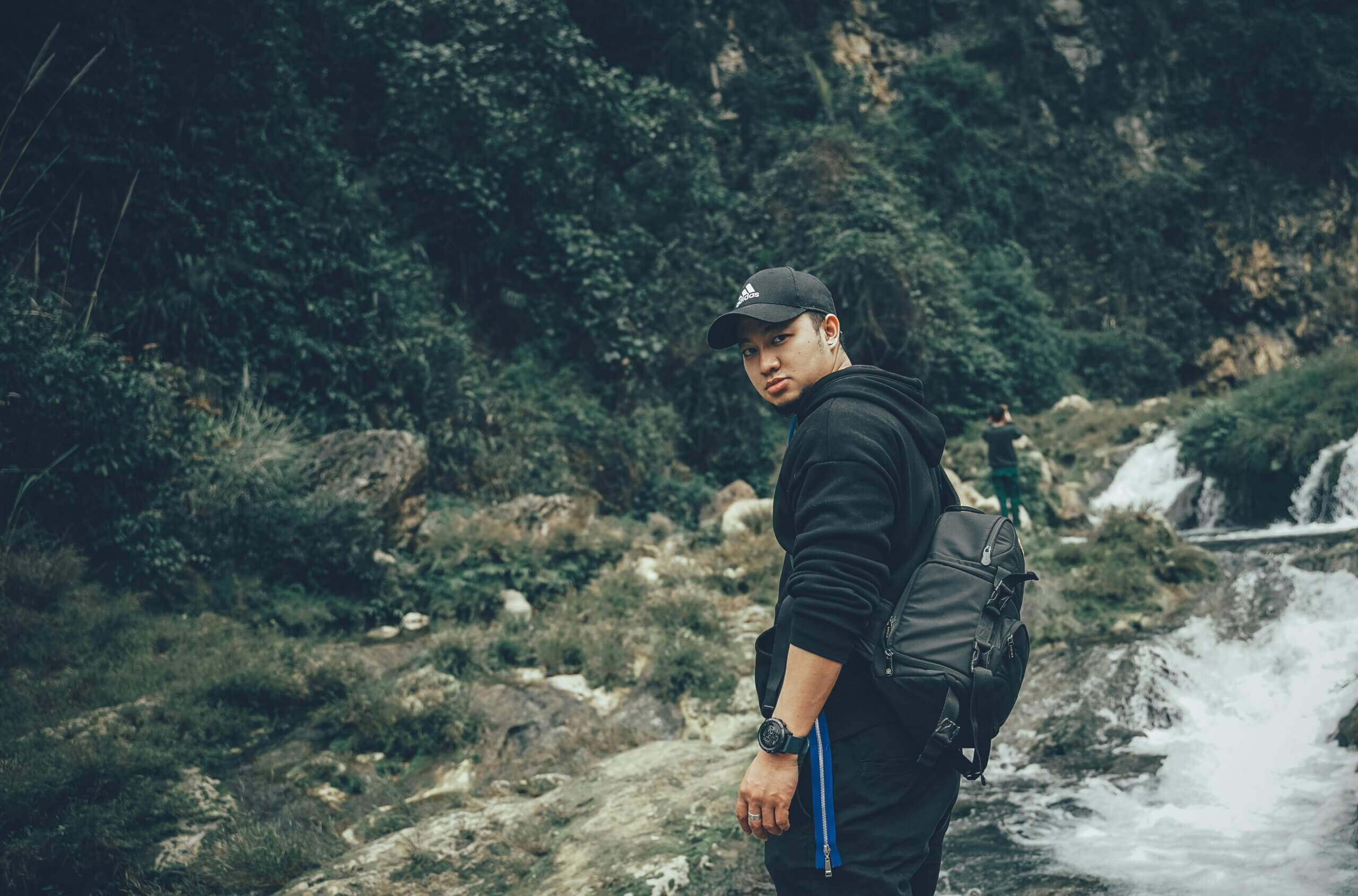 Man wearing black Adidas cap and backpack standing near a waterfall in a forested area.