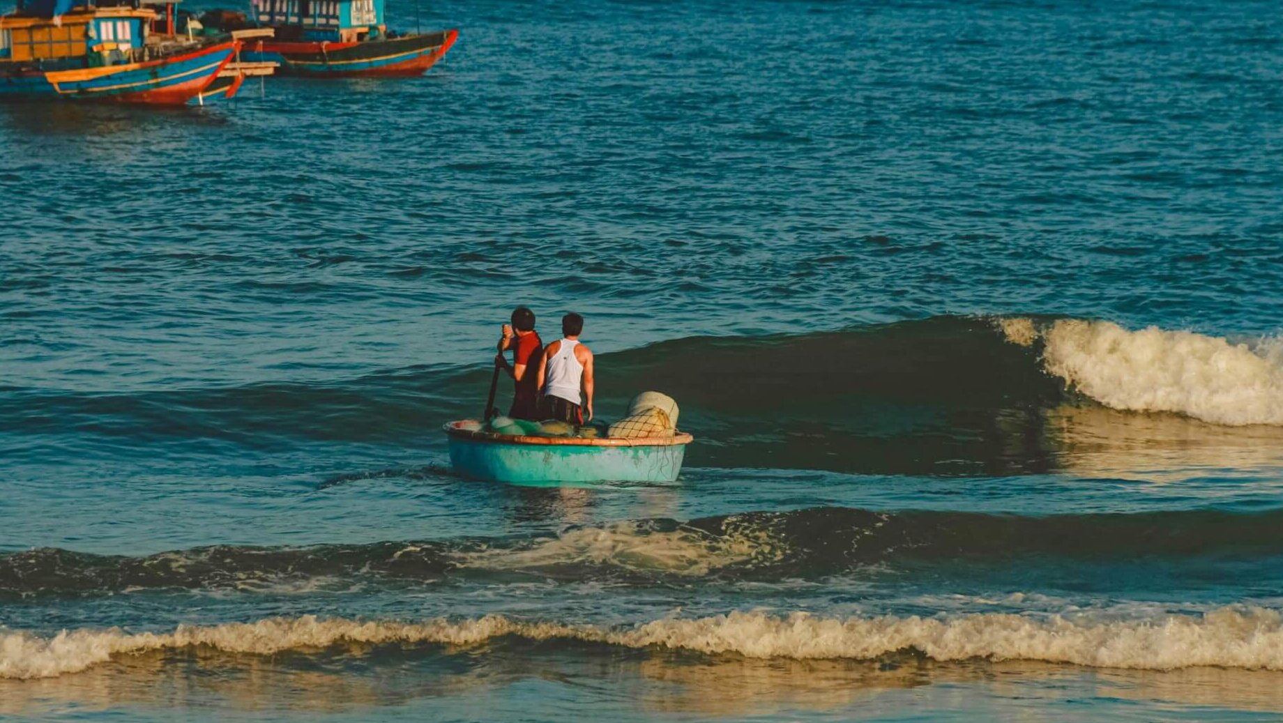 Two people rowing a small round boat toward fishing boats with red flags on the ocean.