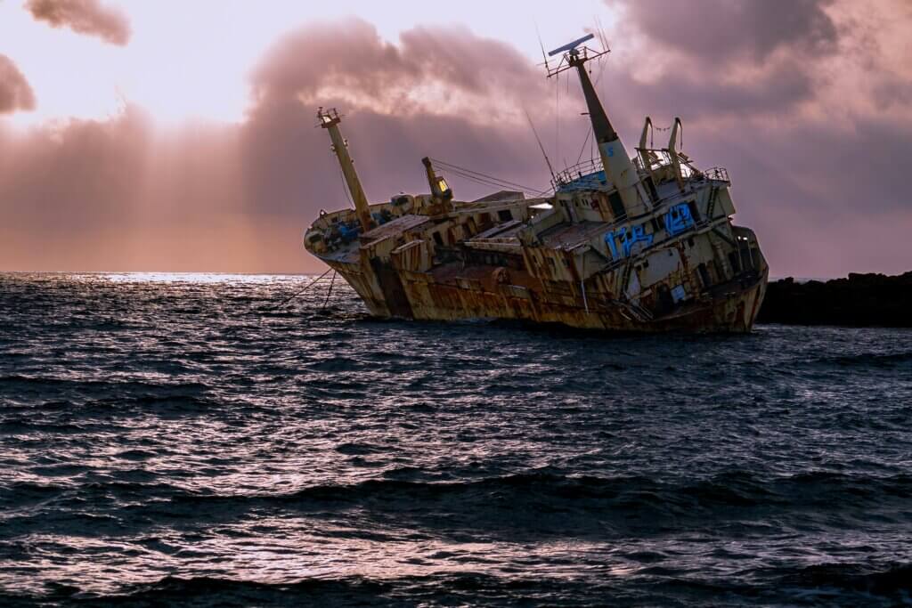 Rusty shipwreck leaning on its side near rocky shore under a cloudy sunset sky