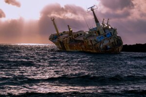 Rusty shipwreck leaning on its side near rocky shore under a cloudy sunset sky