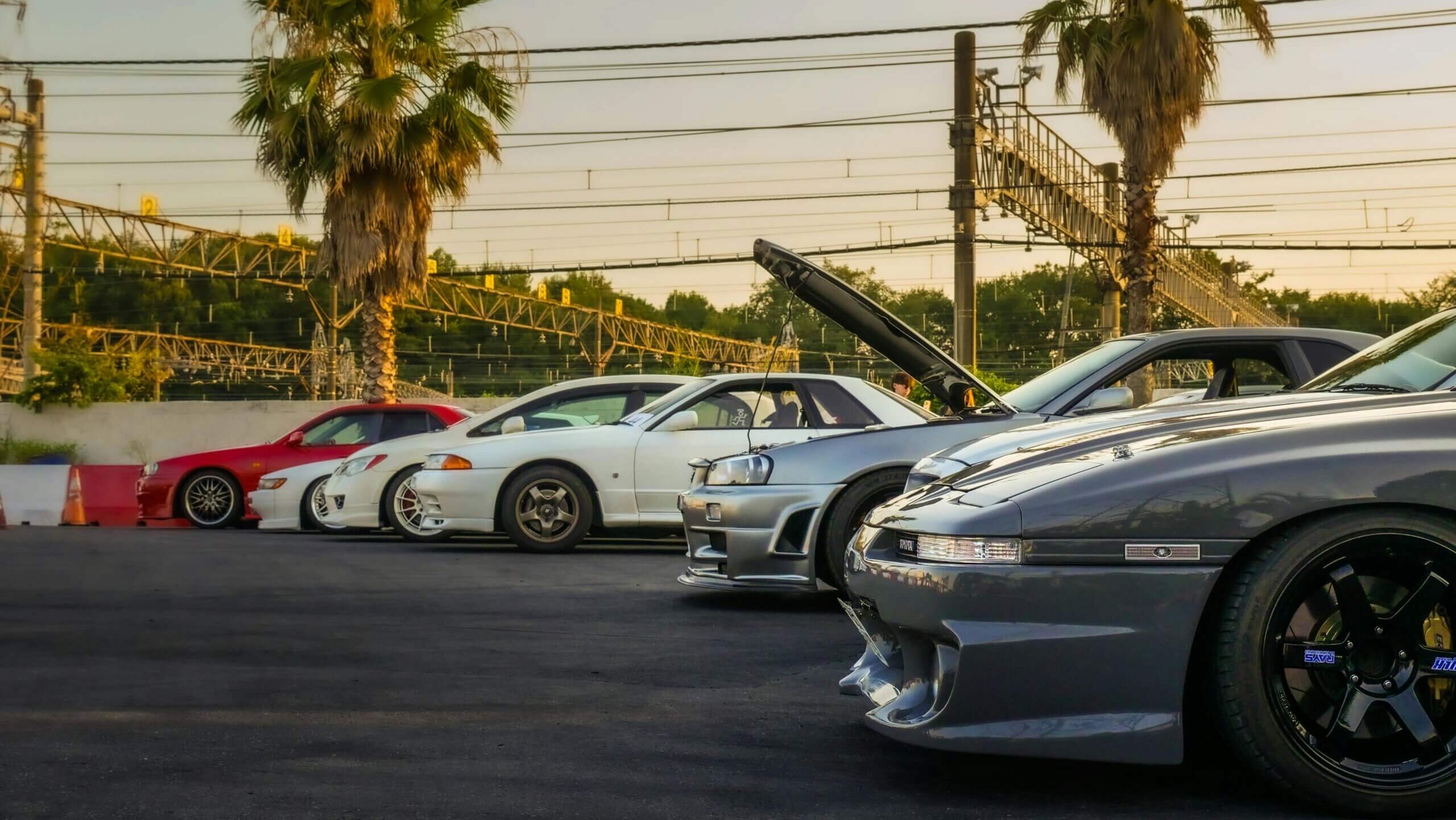 Lineup of modified sports cars including Nissan Skyline models parked near palm trees at sunset