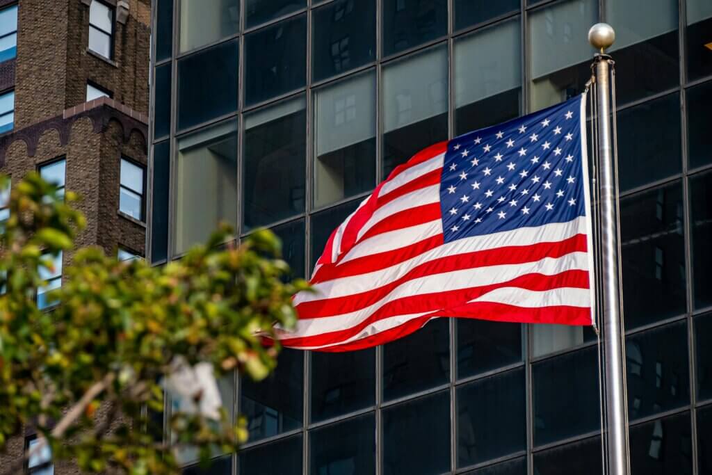 American flag waving on a flagpole in front of a glass building with a brick building in the background