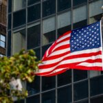 American flag waving on a flagpole in front of a glass building with a brick building in the background