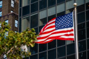 American flag waving on a flagpole in front of a glass building with a brick building in the background