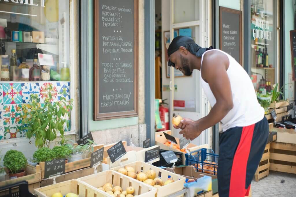 Man in white tank top and black headscarf selecting fruit at outdoor market stall with Portuguese signs.