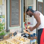 Man in white tank top and black headscarf selecting fruit at outdoor market stall with Portuguese signs.