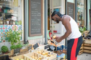 Man in white tank top and black headscarf selecting fruit at outdoor market stall with Portuguese signs.