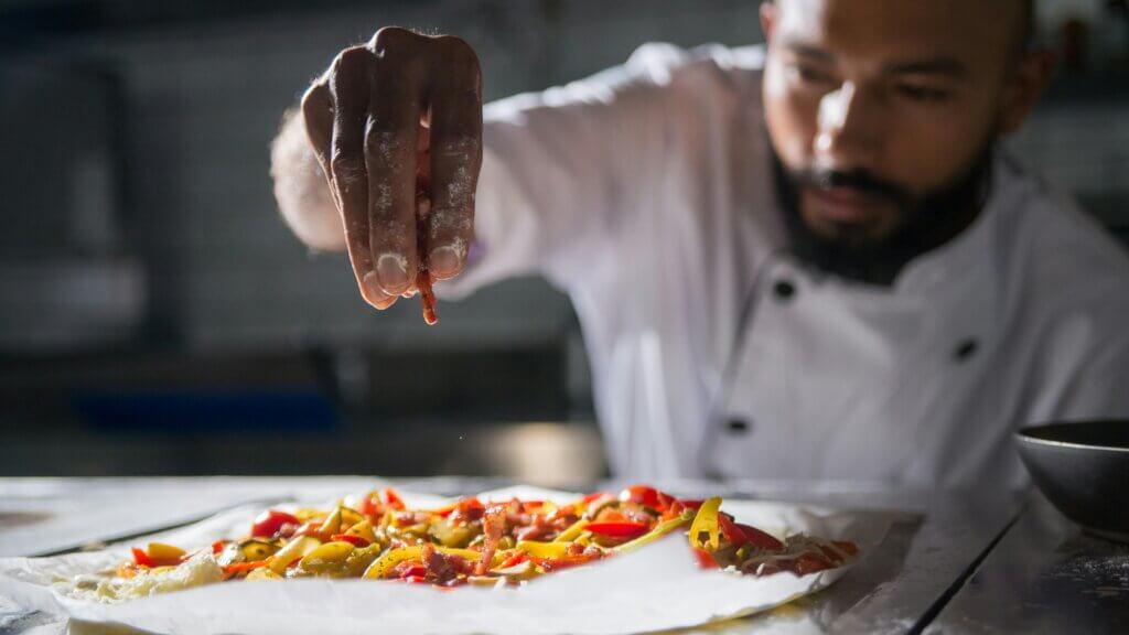 Chef sprinkling toppings on a pizza with colorful vegetables and bacon strips on parchment paper.