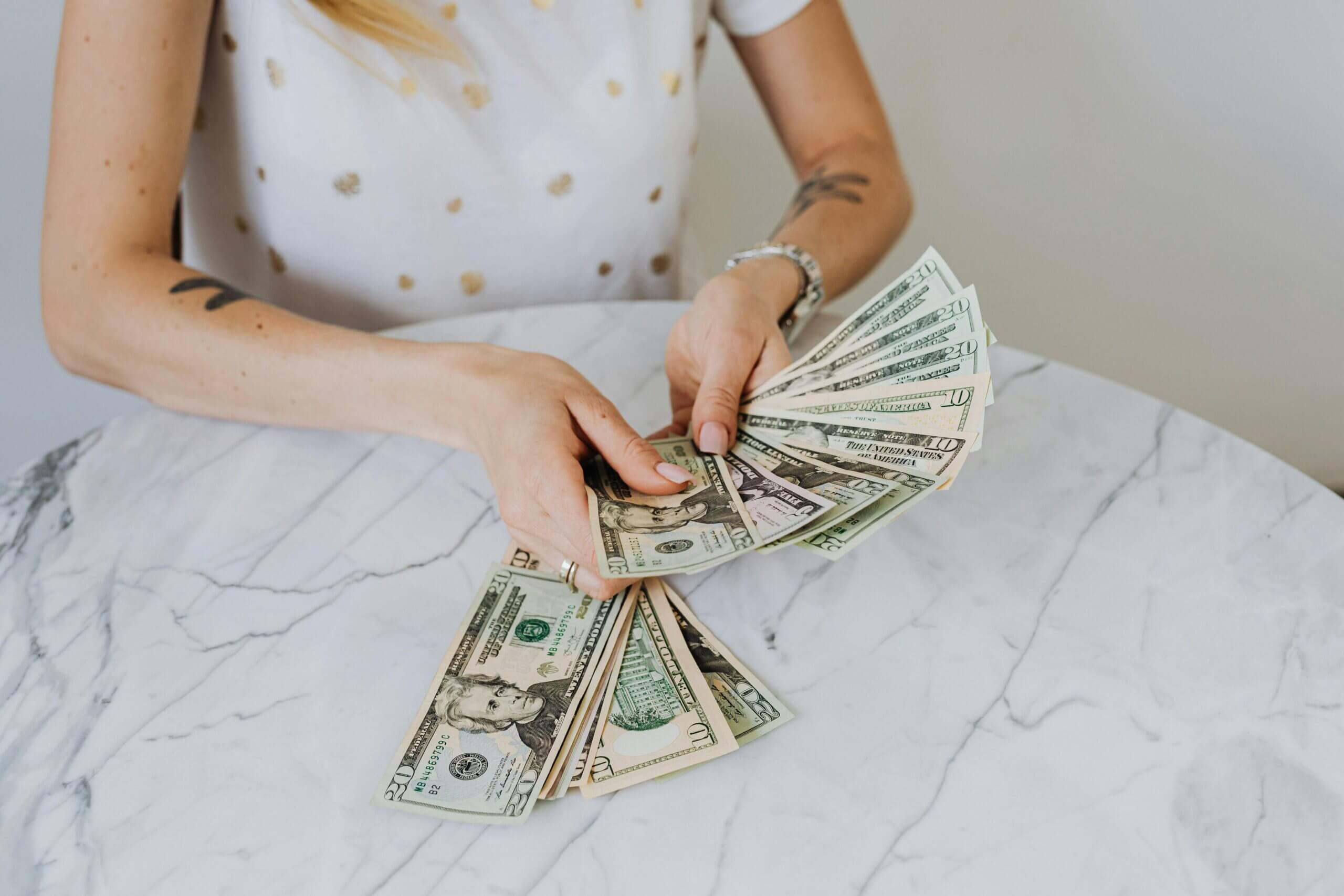 Person with tattoos and a watch holding and fanning out US dollar bills on a marble table