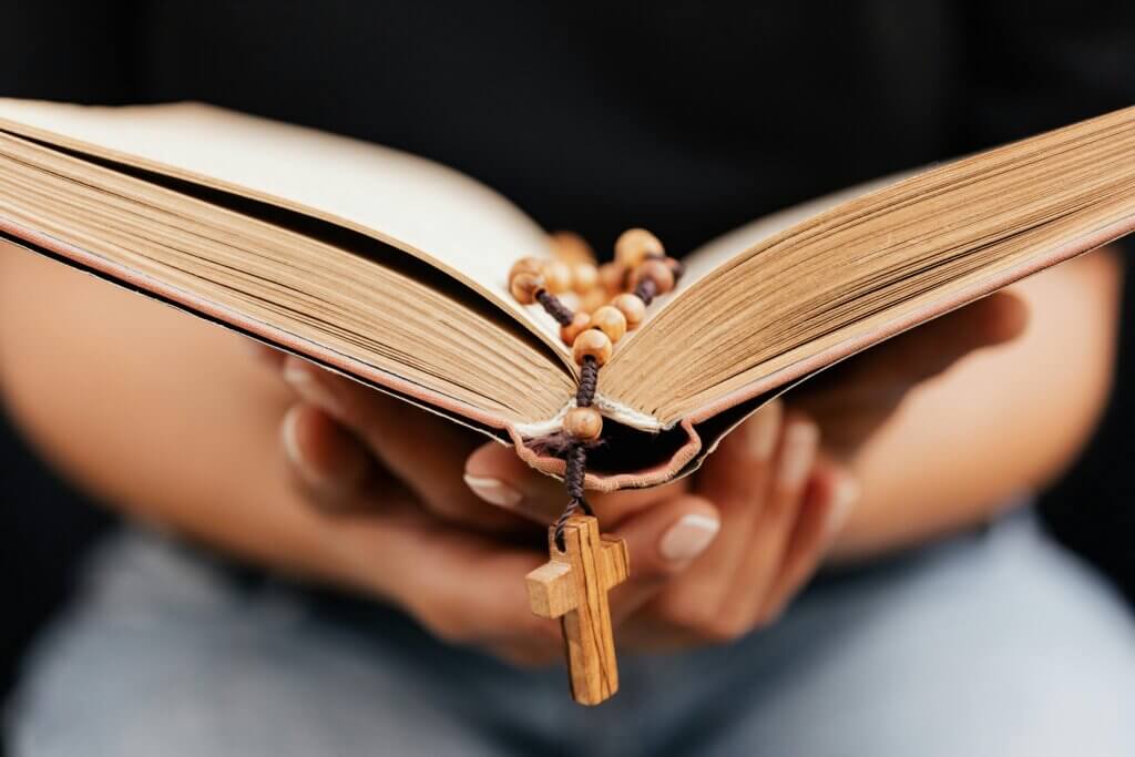 Hands holding an open Bible with a wooden rosary and cross resting on the pages