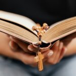 Hands holding an open Bible with a wooden rosary and cross resting on the pages