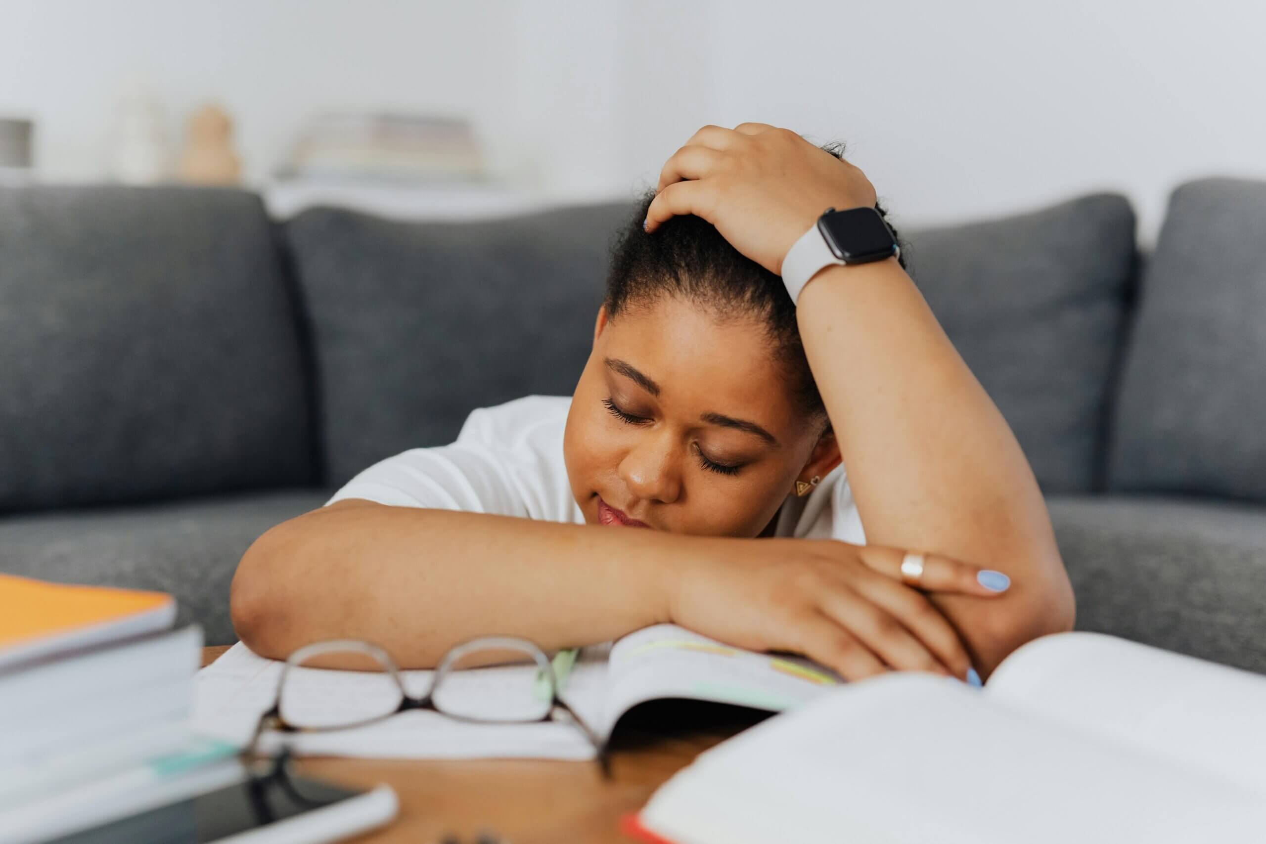 Tired young woman resting her head on her arms over an open book with glasses and notebooks nearby.