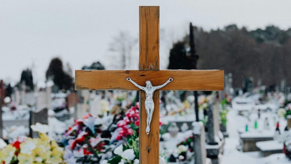 Wooden crucifix with metal figure of Jesus in a snowy cemetery with colorful flowers on graves