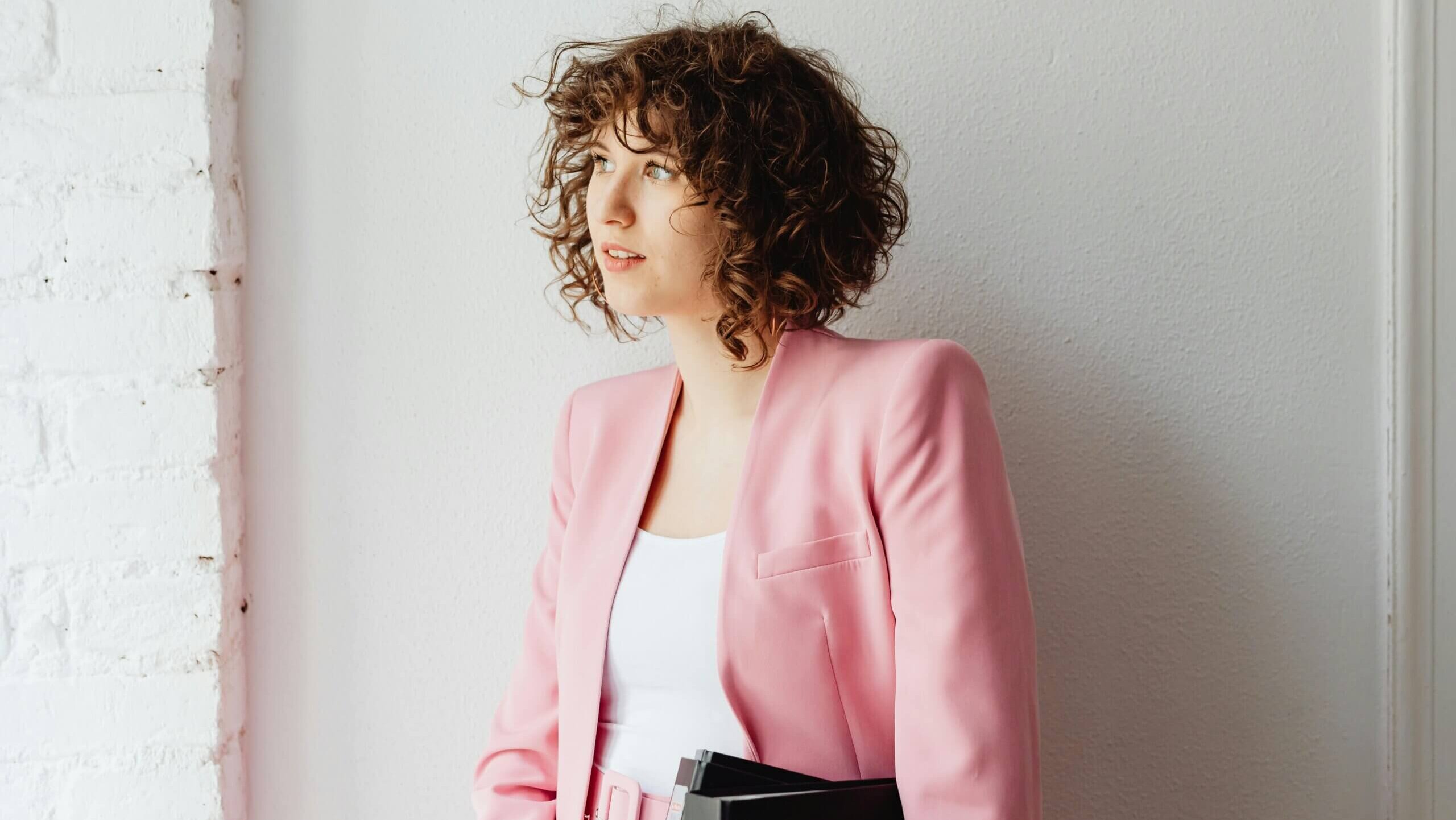 Young woman with curly hair wearing a pink blazer and holding black folders, looking to the side against a white wall.