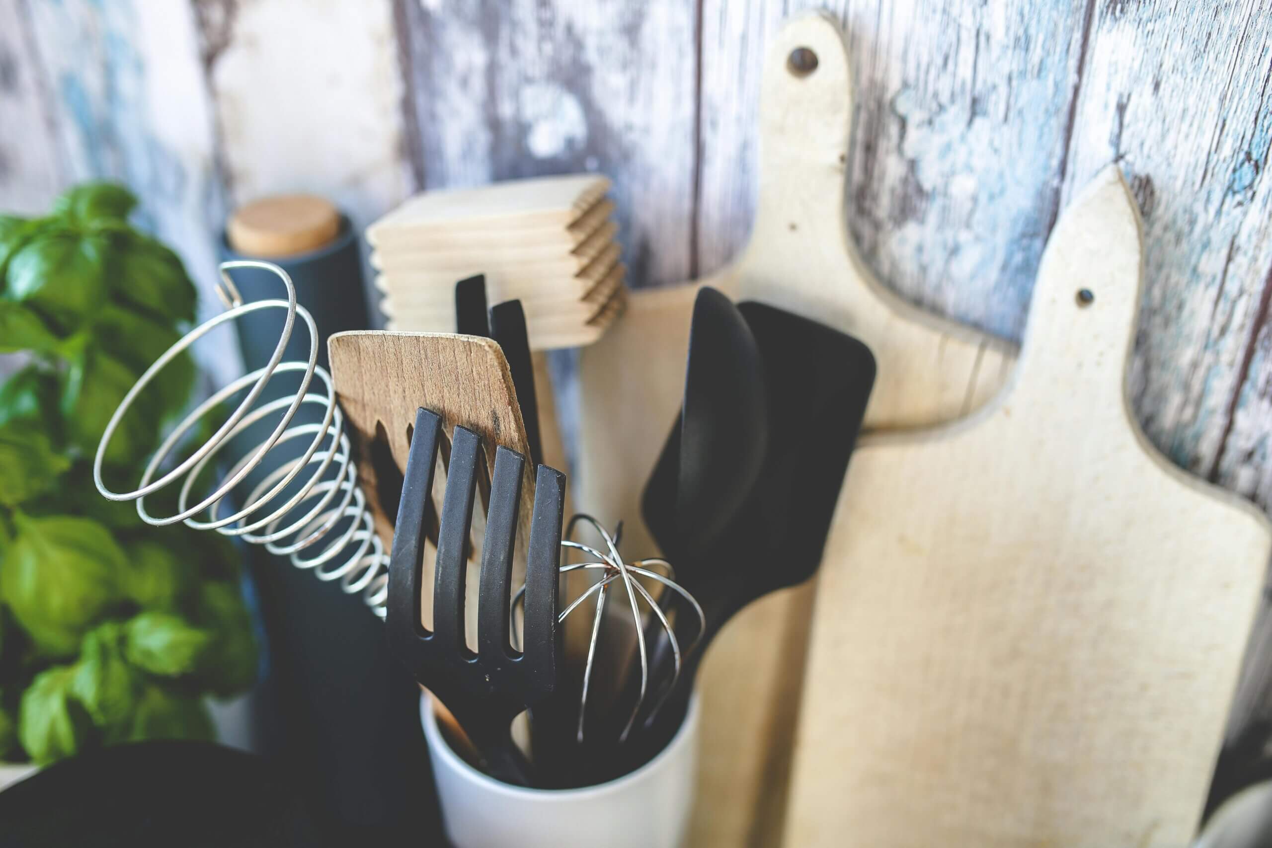 Kitchen utensils including whisks, spatulas, and wooden spoons in a white holder with wooden cutting boards in the background