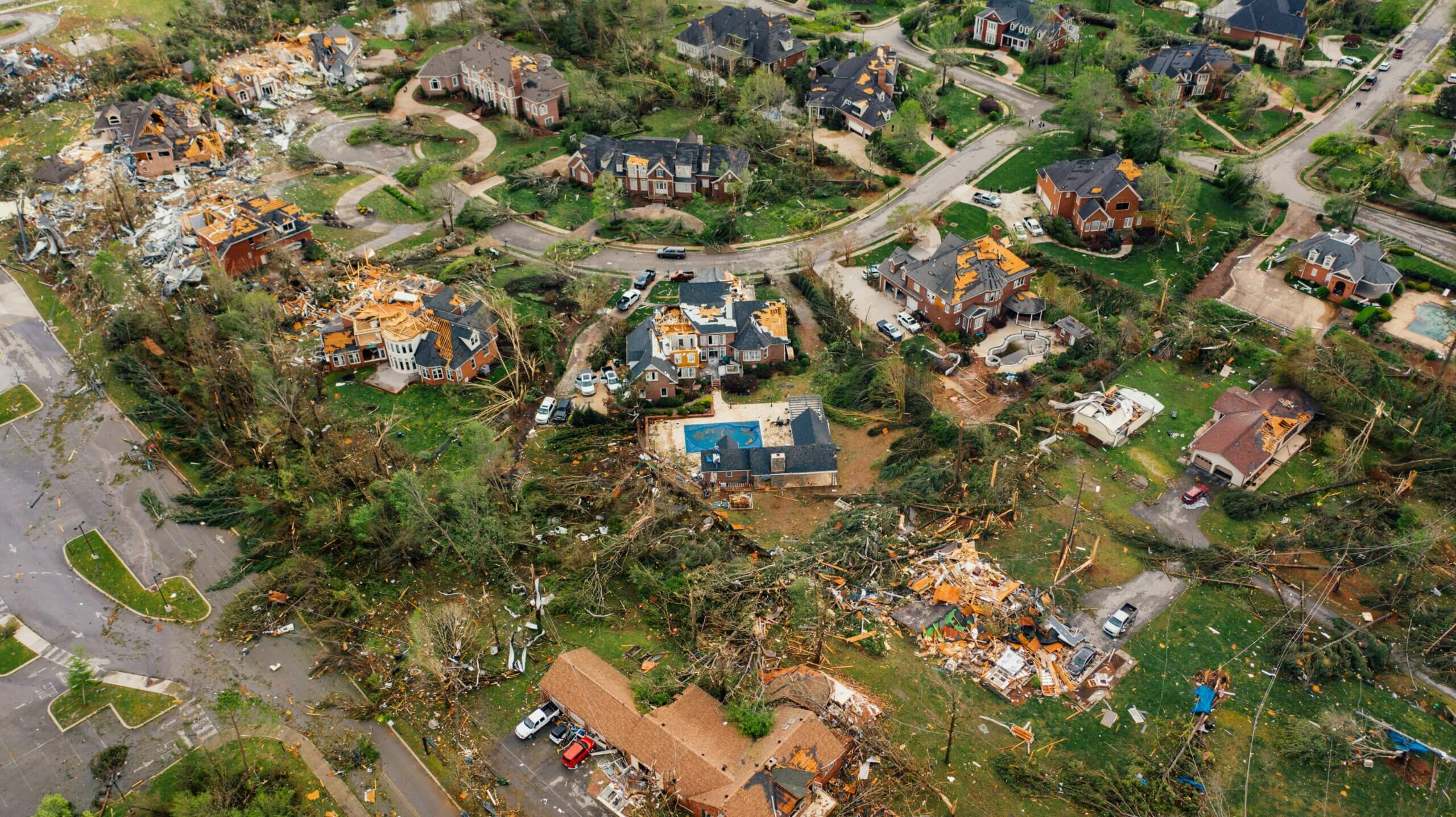 Aerial view of a residential neighborhood with multiple homes severely damaged by a tornado, showing debris and fallen trees.