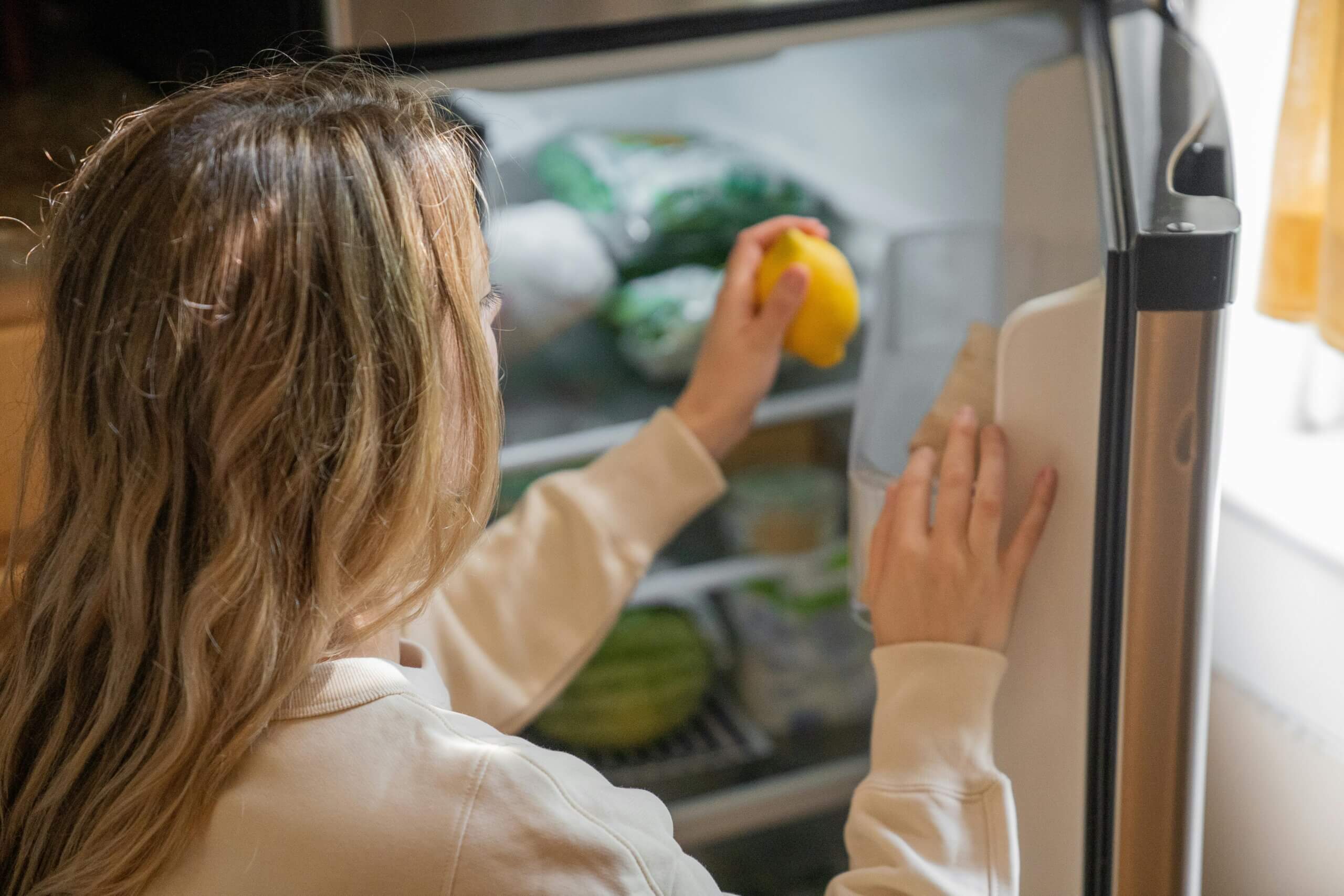 Person in beige sweatshirt taking a lemon from an open refrigerator door.