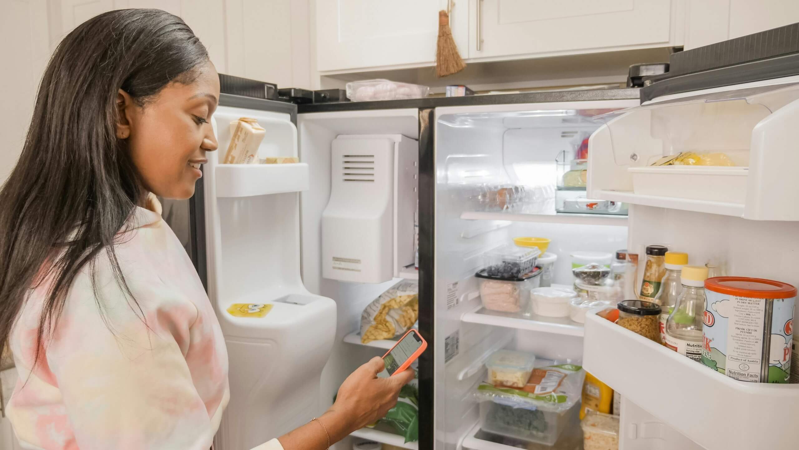Woman in a tie-dye hoodie checking her phone while standing next to an open refrigerator filled with food and condiments.
