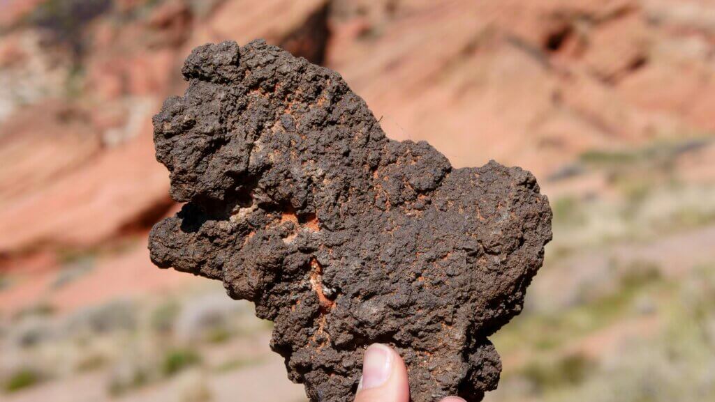 Hand holding a rough, dark volcanic rock with a blurred desert landscape in the background.