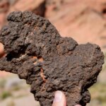 Hand holding a rough, dark volcanic rock with a blurred desert landscape in the background.