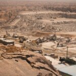 Open-pit quarry with heavy machinery and conveyor belts processing rocks in a dry, rocky landscape.
